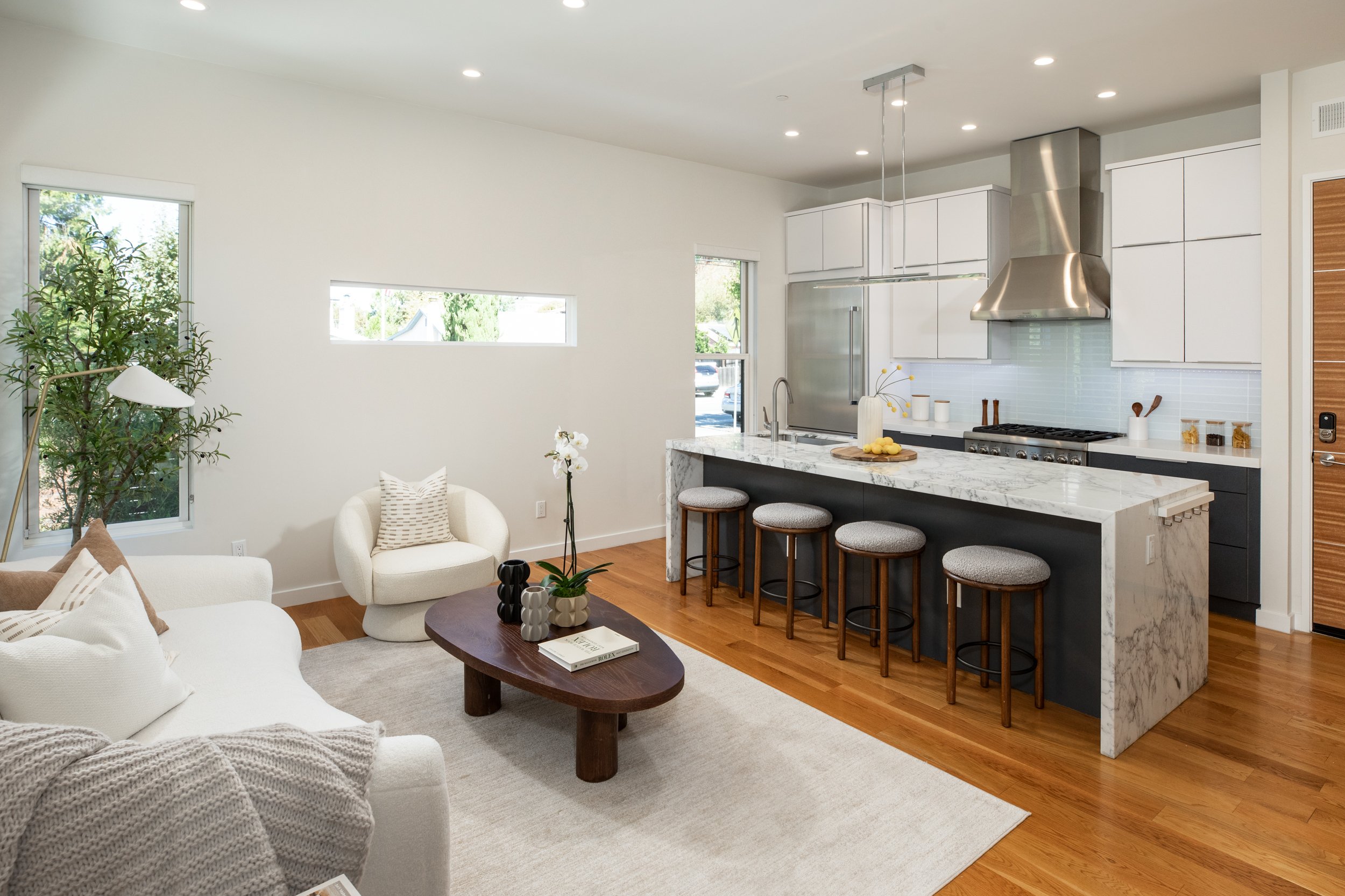 A modern open-concept living room and kitchen with white walls, hardwood floors, and large windows. The living room has a white sofa with throw pillows, a round wooden coffee table with decorative vases, and a light-colored armchair. The kitchen feat