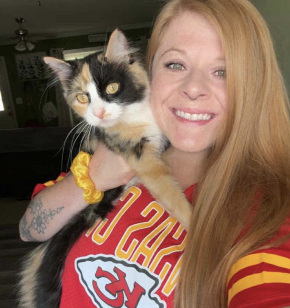 picture of a woman with a Kansas City Cheifs jersey smiling while holding a brown and black cat