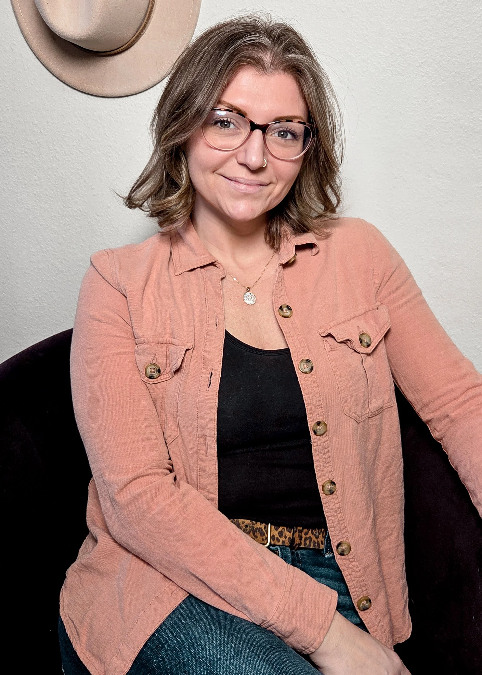 A woman with shoulder-length brown hair, wearing glasses, a nose ring, a black top, a light pink jacket, and blue jeans, sitting against a plain light-colored wall. There is a hat hanging on the wall behind her.