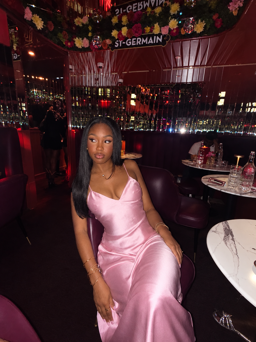 A woman in a pink satin dress sitting in a dimly lit restaurant or lounge with city lights visible through the window behind her. Decor includes flowers and shiny hanging ornaments.