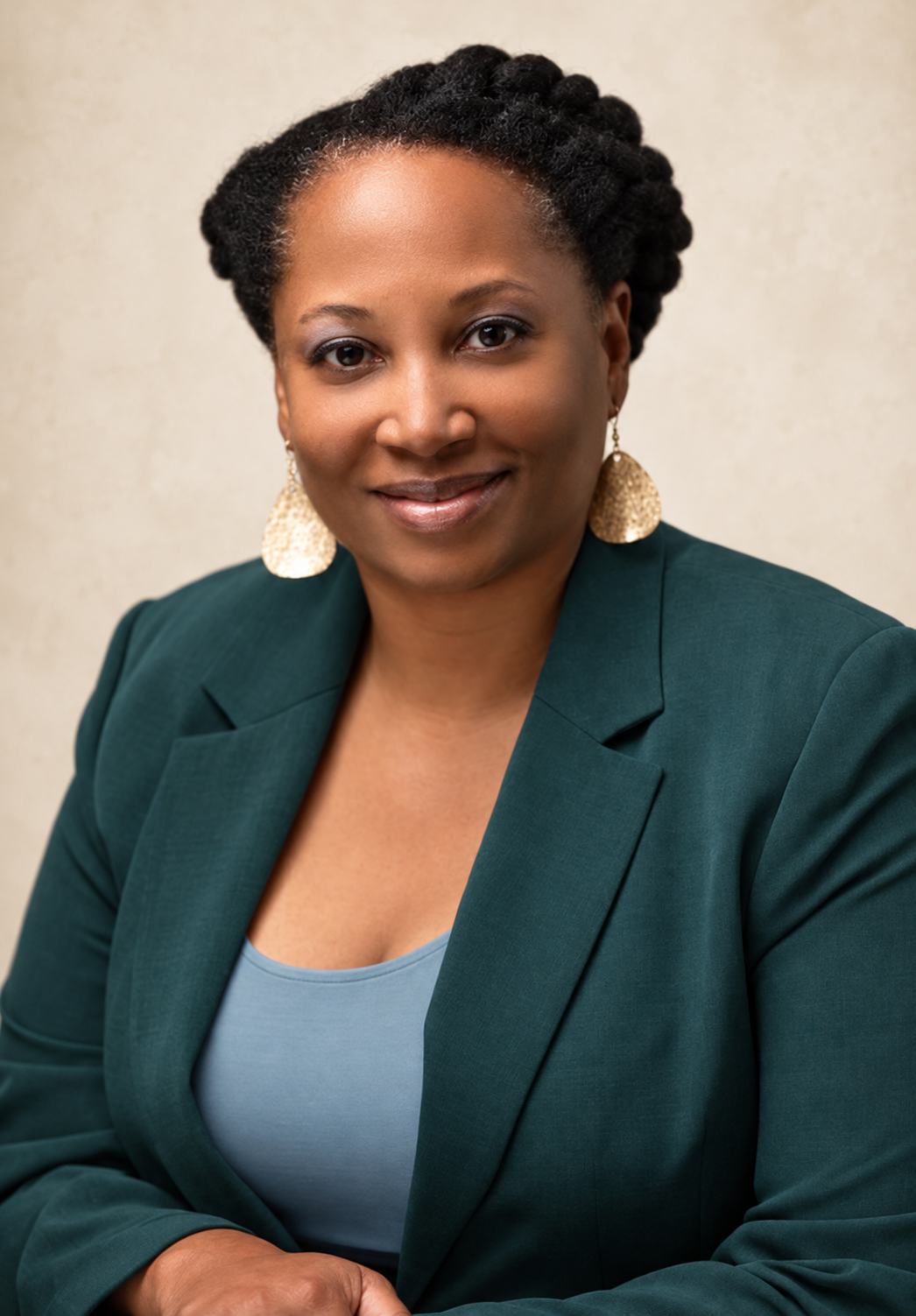 A woman with short natural hair, wearing a teal blazer, grey top, and gold earrings, smiling in front of a neutral background.
