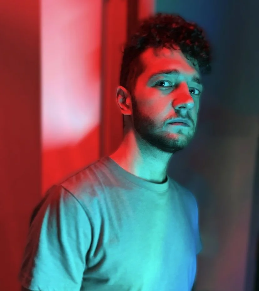 Young man with curly hair and facial hair standing against a wall, lit with red and blue colored lights.