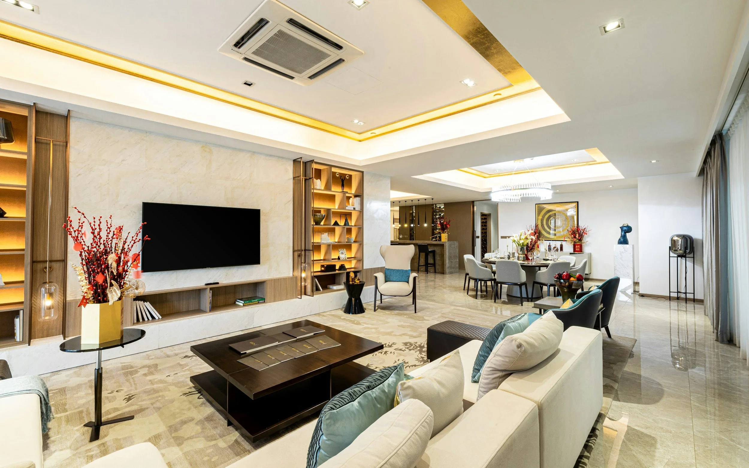 A spacious modern living room with a cream-colored sofa, a dark wood coffee table, and a TV mounted on a white marble wall. The room features built-in shelves with decorative items, a dining area with a round table and white chairs, and elegant lighting fixtures on the ceiling. There are large windows with curtains, and the decor includes flowers and art pieces.