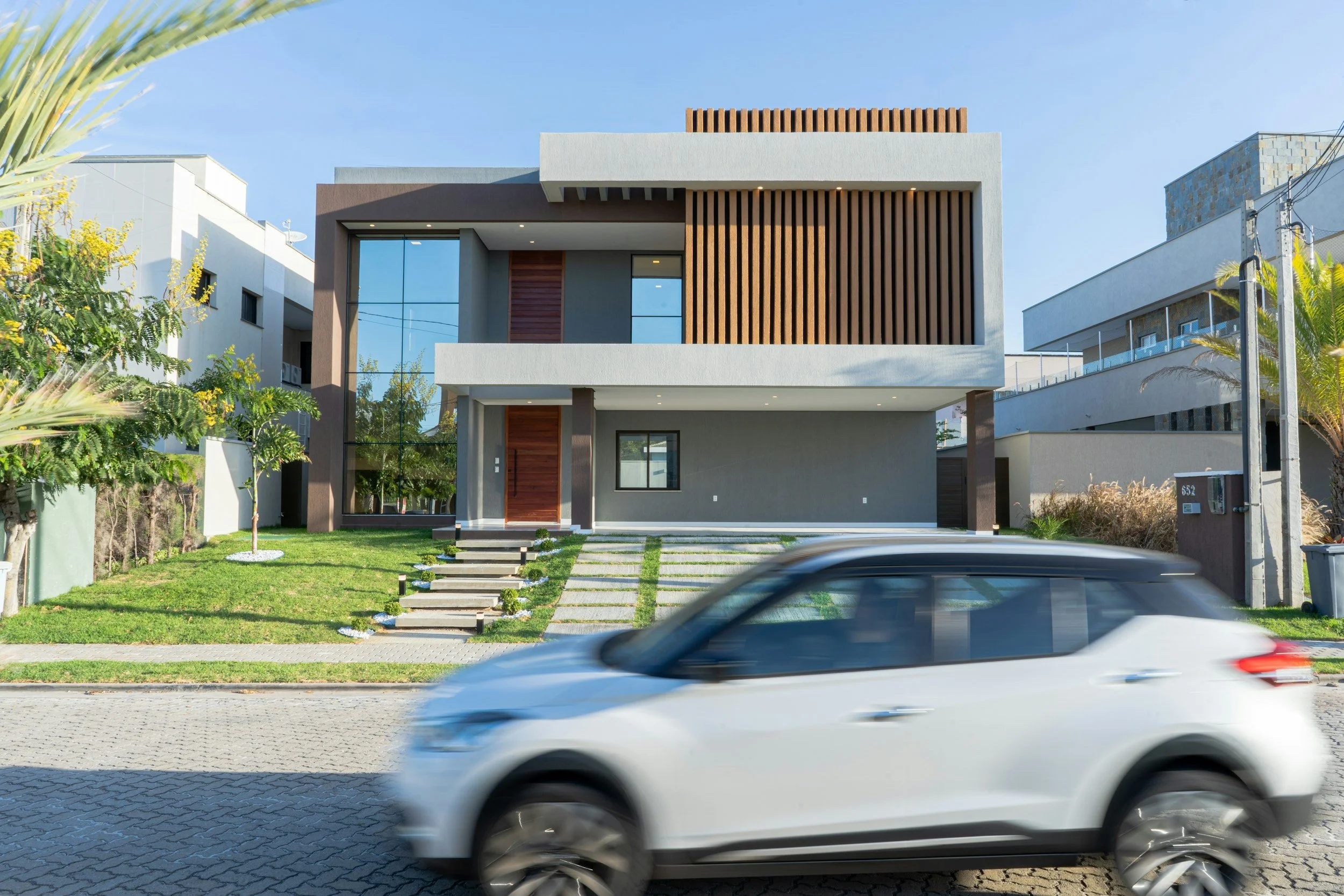 Modern multi-story house with gray and brown exterior, large glass windows, wooden accents, and a landscaped front yard with a pathway. A blurred white car is passing in front.