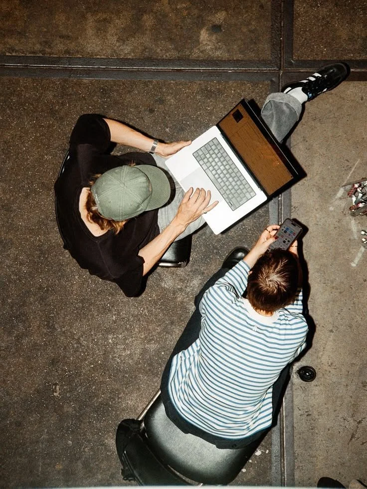 Two people are sitting on chairs at a table, viewed from above. The person on the left is wearing a green cap and black shirt, using a laptop, while the person on the right has brown hair and a striped shirt, holding a smartphone.