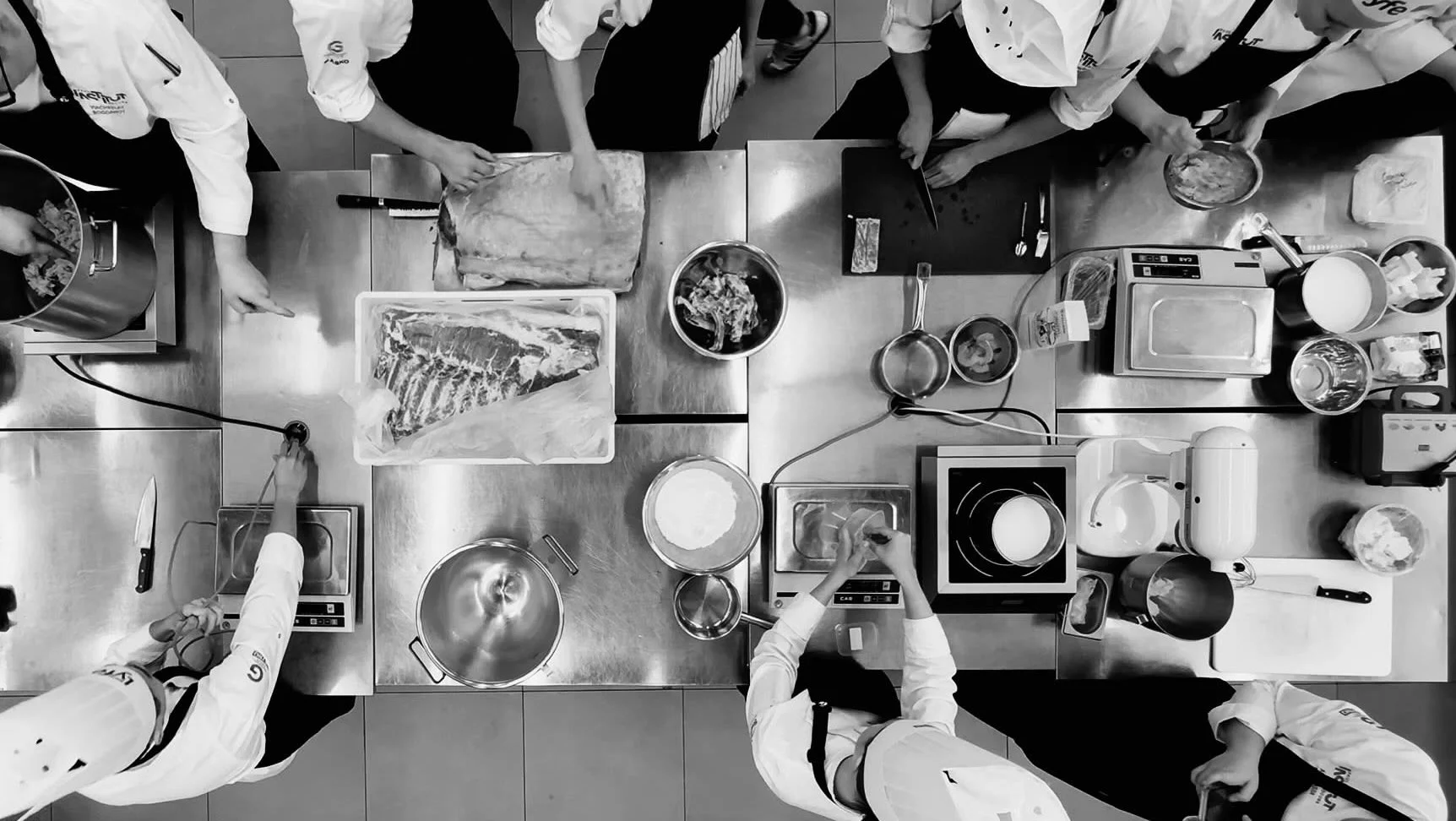 Chefs in a professional kitchen preparing meat and ingredients for cooking, with various utensils, pots, and kitchen appliances on stainless steel countertops.