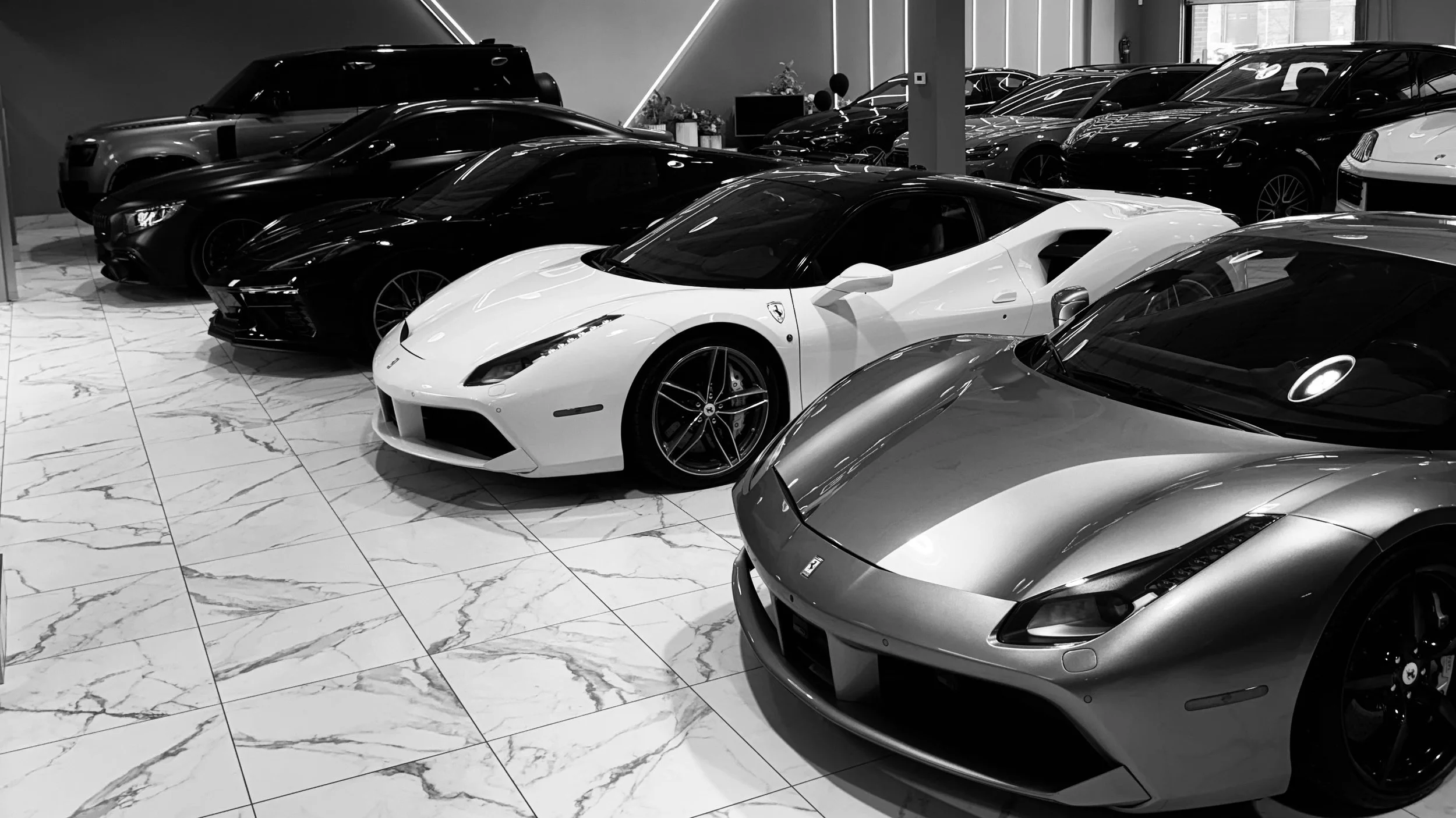 Multiple luxury cars, including a white sports car in the foreground, parked inside a showroom with a marble floor and modern interior design.