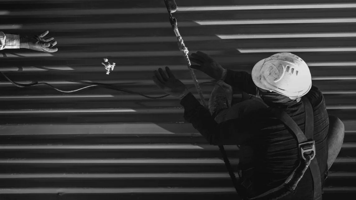 A worker in a safety helmet and gloves is installing or repairing a corrugated metal roof using a harness and safety ropes.