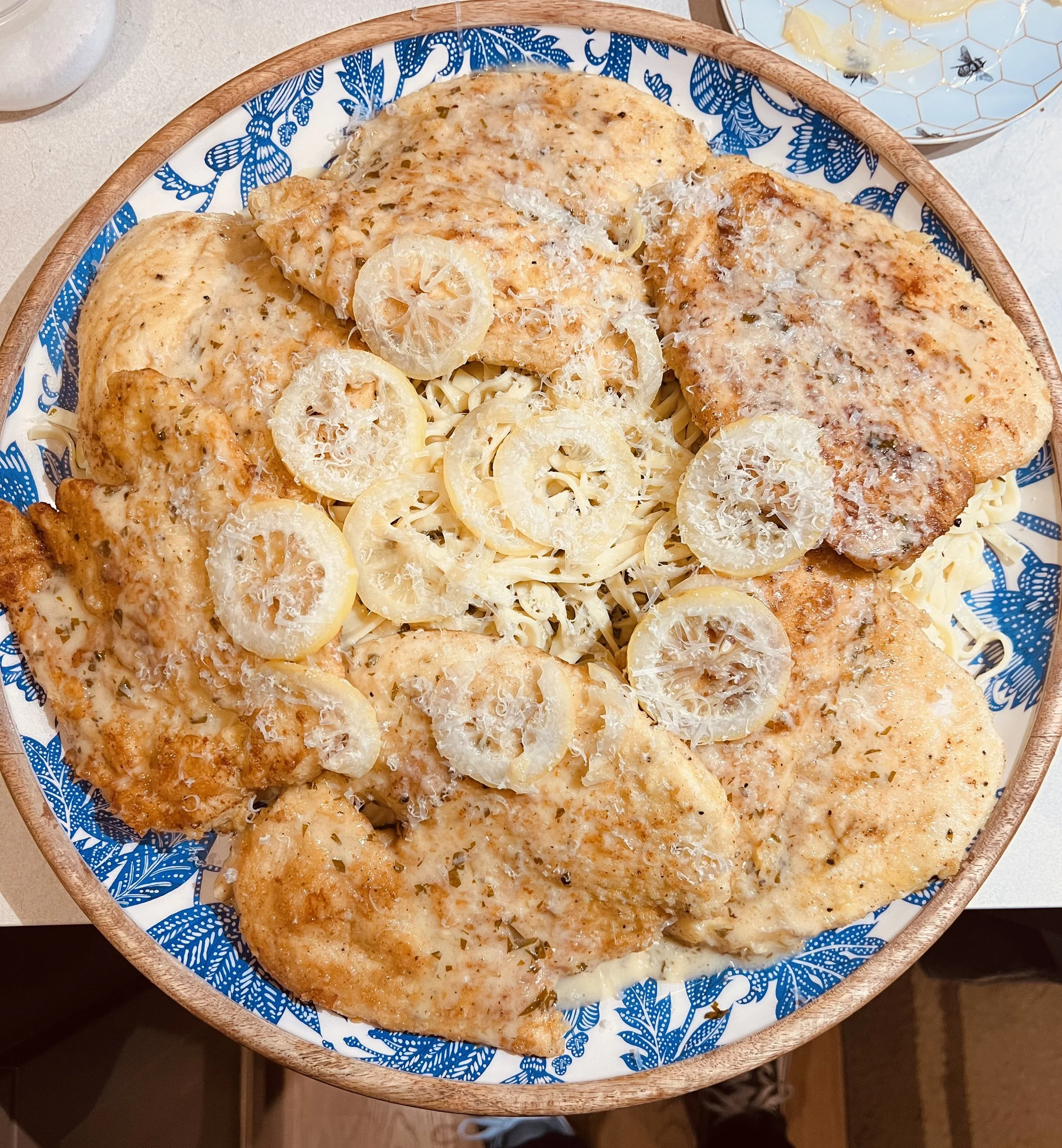Plate of cooked chicken breast topped with grated cheese, served with sliced lemon and pasta, on a decorative blue and white plate.