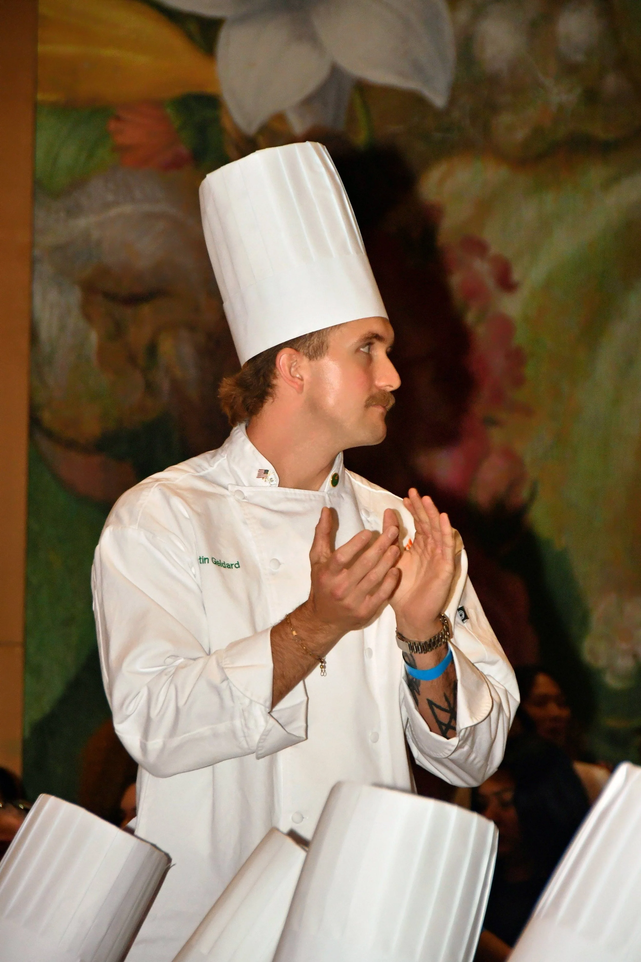 A male chef wearing a traditional white chef's coat and tall chef hat, standing in front of a colorful mural, clapping his hands.