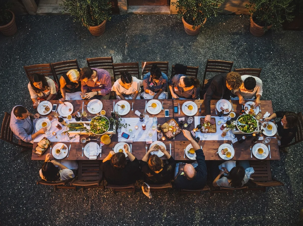 Overhead view of a large group of people sitting at a rectangular wooden table outdoors, sharing a meal with plates, glasses, and decorative items.