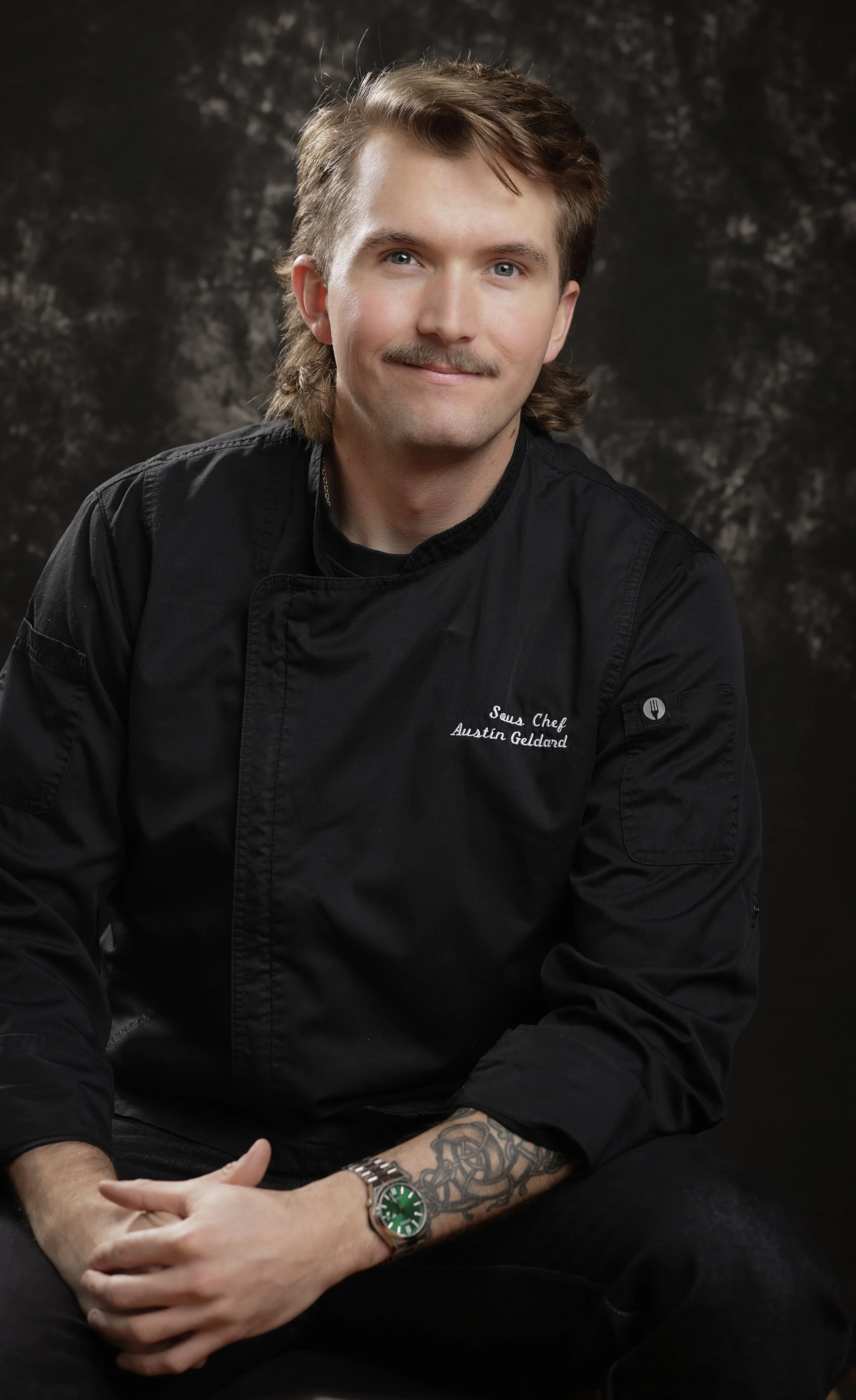 Portrait of a man with light skin, brown hair, a mustache, dressed in a black chef's jacket with ''Sous Chef Austin Gelcard'' embroidered on it, wearing a silver watch with a green face and tattoos on his left forearm, sitting against a dark textured background.