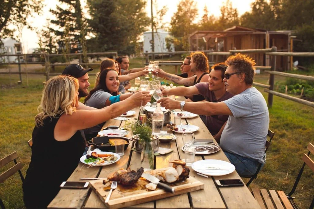 A group of people, sitting around a long outdoor table in a backyard, toasting their glasses during sunset.