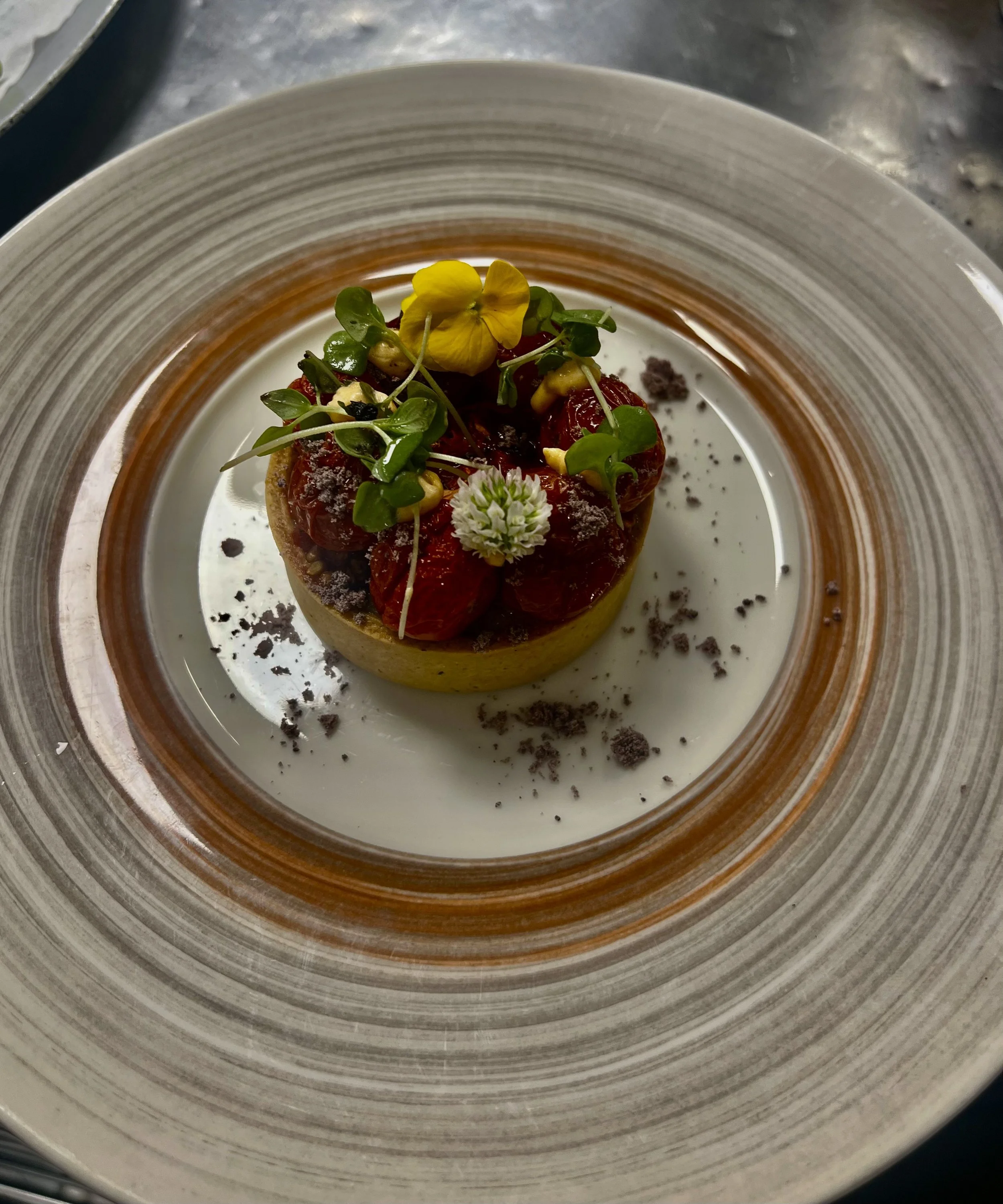 Close-up of a plated gourmet dish with a round base topped with tomato slices, microgreens, edible flowers, and garnished with black and white crumbs, served on a striped ceramic plate.