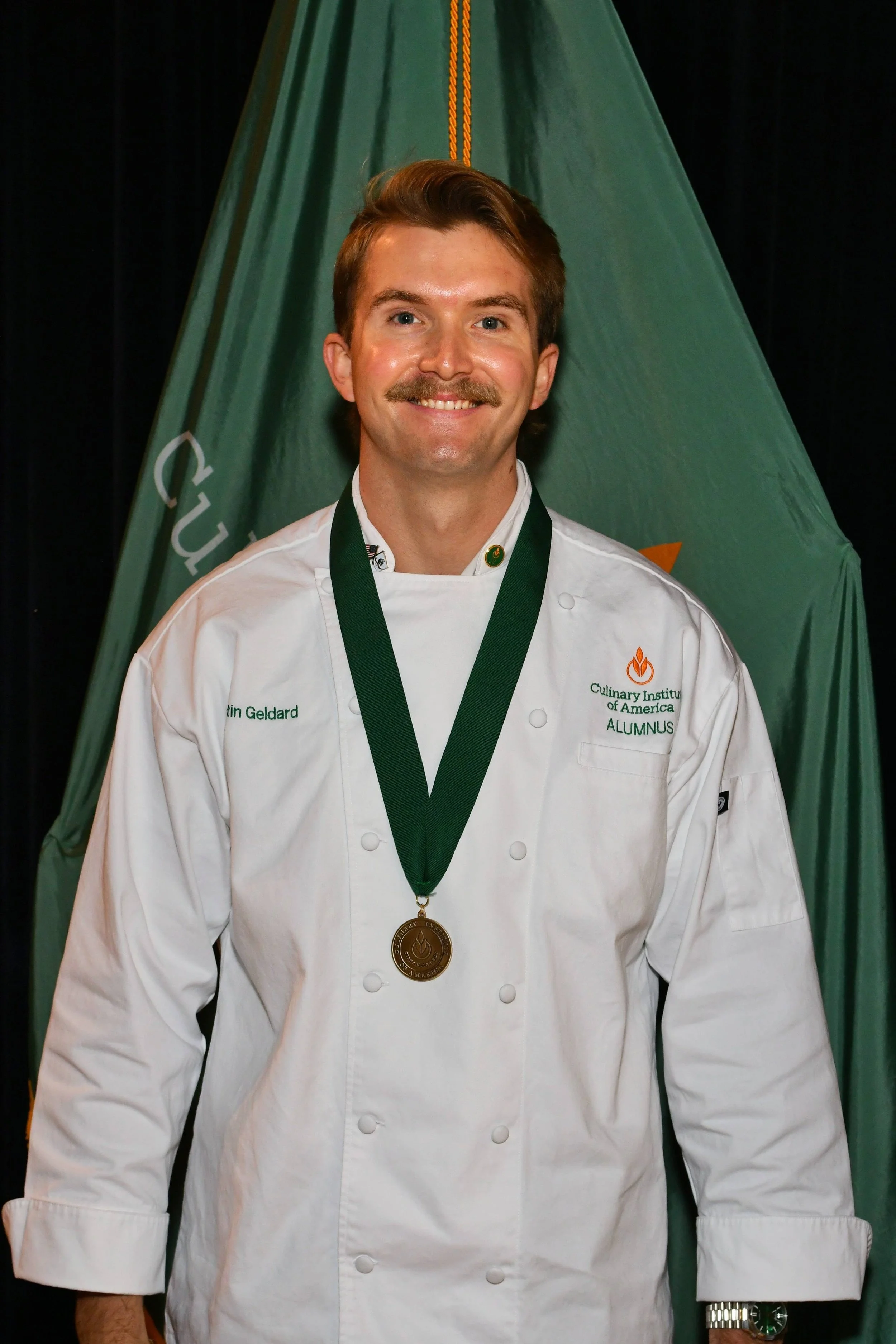 A man smiling in a white chef's coat with a gold medal around his neck, standing in front of a green flag with orange and white lettering. He is wearing a wristwatch and has a mustache.