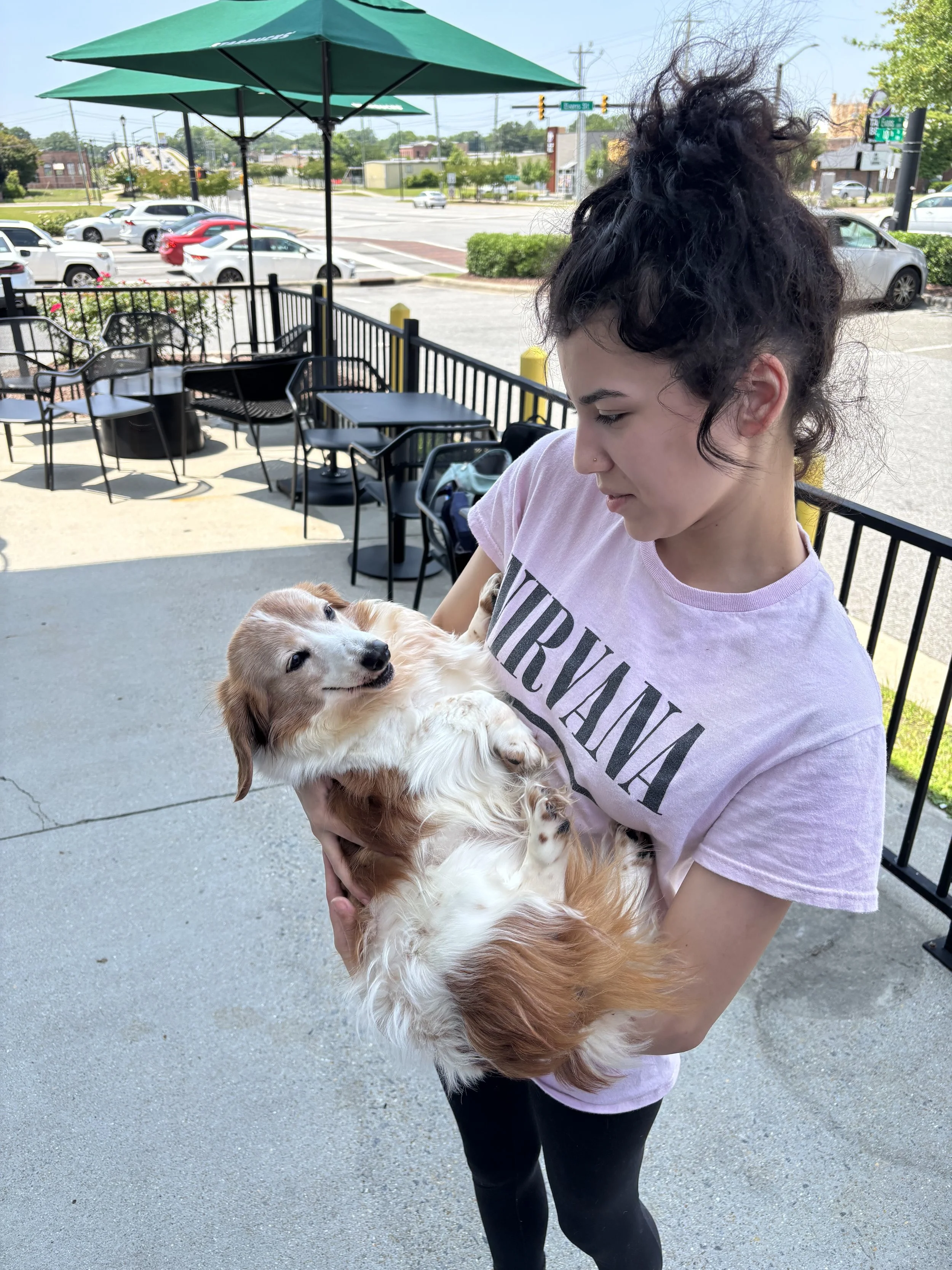 A woman with curly dark hair holding a puppy in an outdoor seating area on a bright day. The woman is wearing a light pink t-shirt with the word 'VIRGINIA' printed on it and black leggings. The background shows a parking lot, parked cars, green umbrellas, and a street intersection.