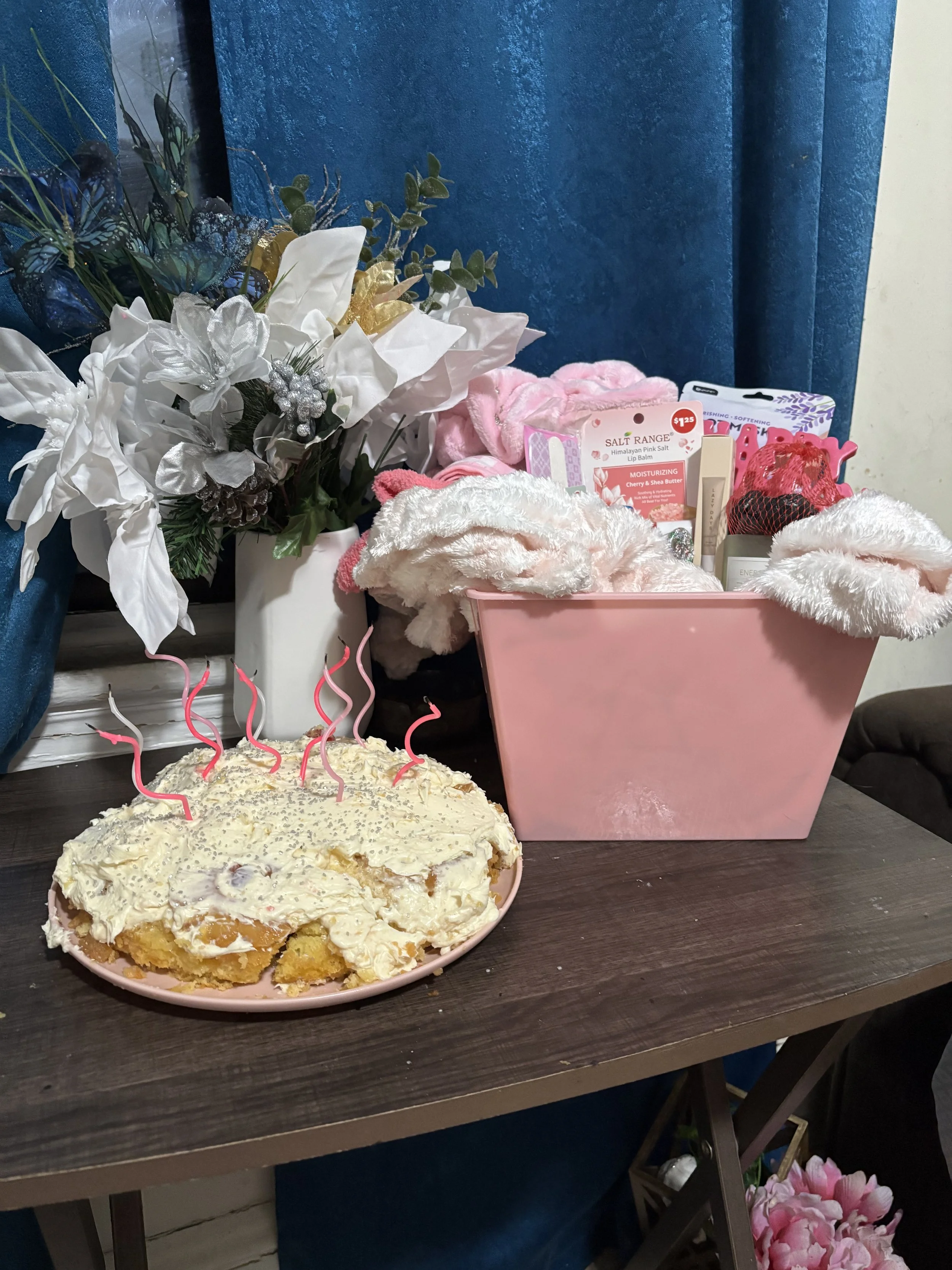 A partially eaten peach-colored cake with white frosting and pink candles on a pink plate on a wooden table. In the background, there is a white floral arrangement with artificial flowers and green leaves, pink plush slippers, and various personal care items in pink packaging. A blue curtain and part of a yellow wall are behind the table.