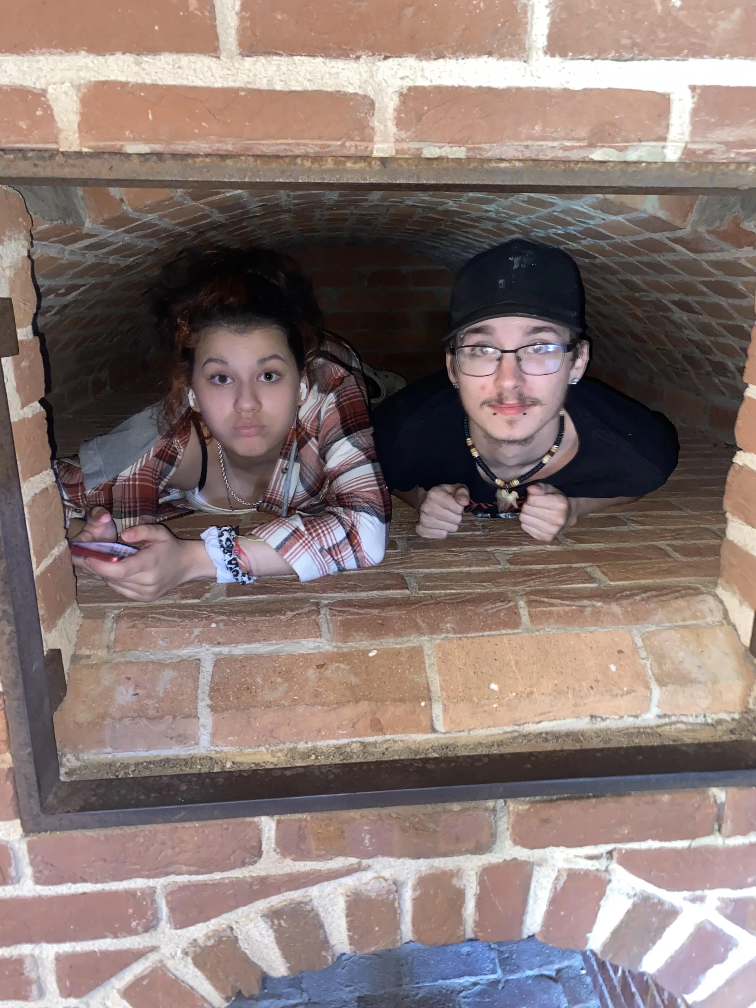 Two people lying inside a brick oven, looking out of the opening at the camera.