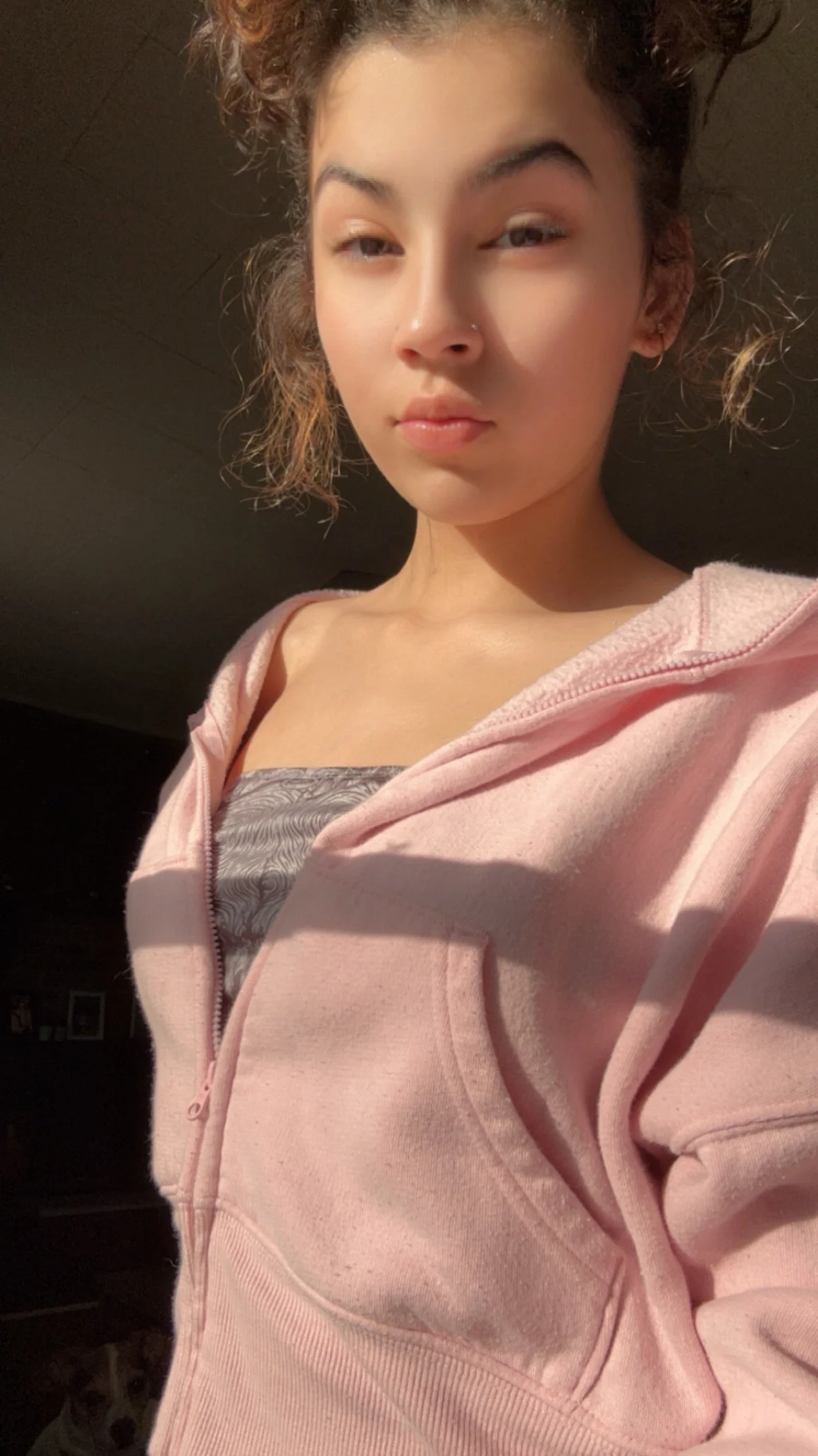 A young woman with curly hair and a nose piercing is wearing a pink hoodie over a patterned top. She is taking a selfie indoors with soft lighting.