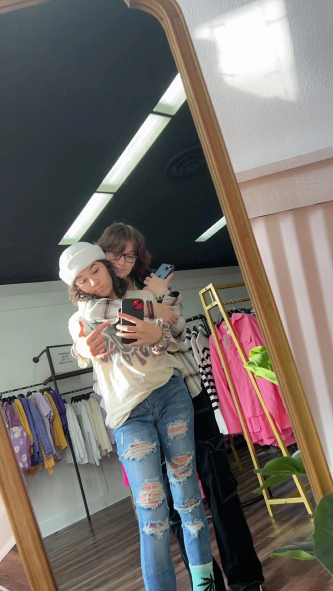 Makayla and her sister taking a mirror selfie in a clothing store, one wearing a white hat and distressed jeans, the other with glasses and a black shirt, standing close together, with various clothing racks and pink bags in the background.
