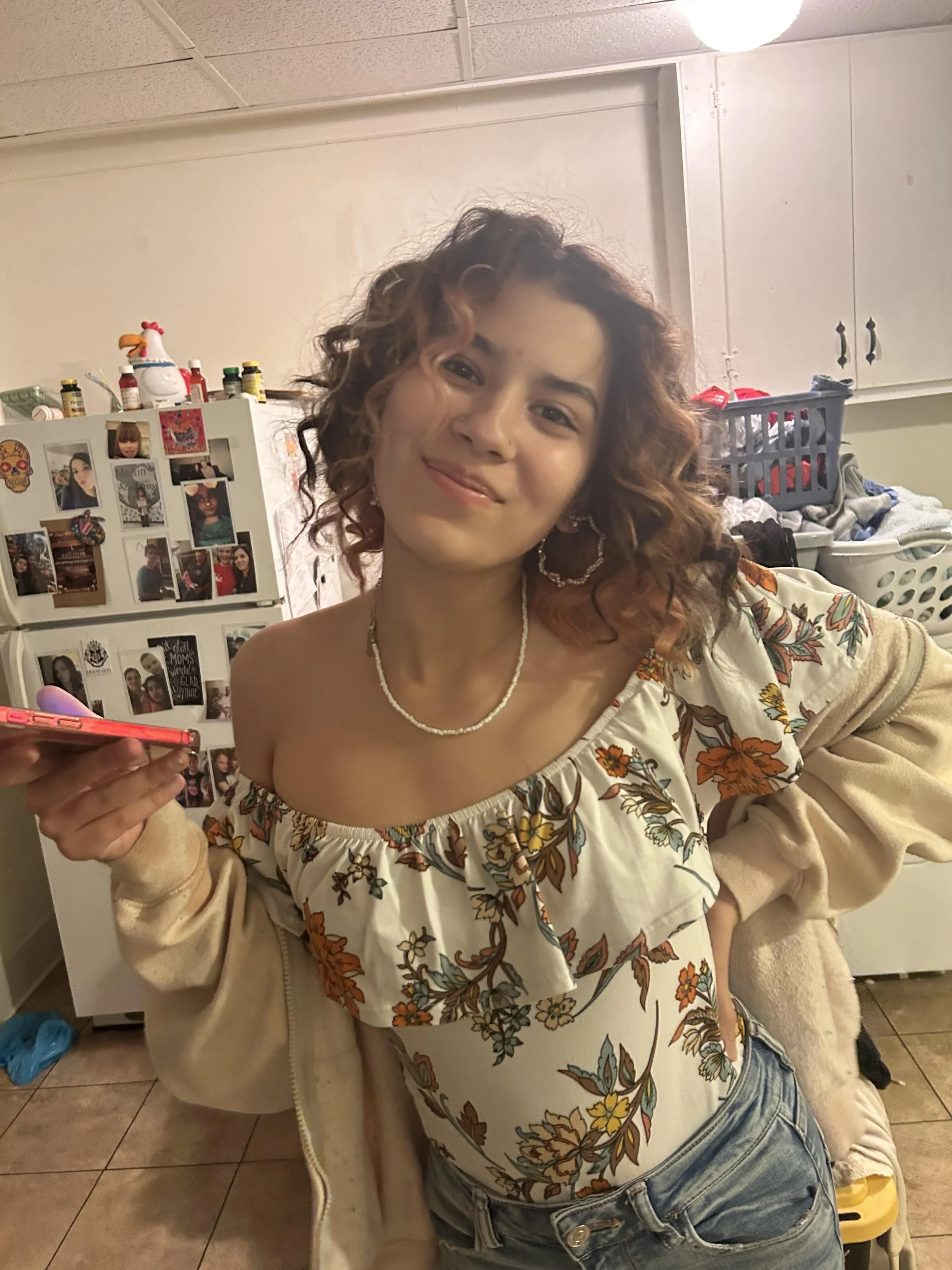 Makayla with curly hair, wearing a floral off-shoulder top and a beige jacket, smiling and taking a selfie in a kitchen. There are laundry baskets and a refrigerator with photos and magnets in the background.