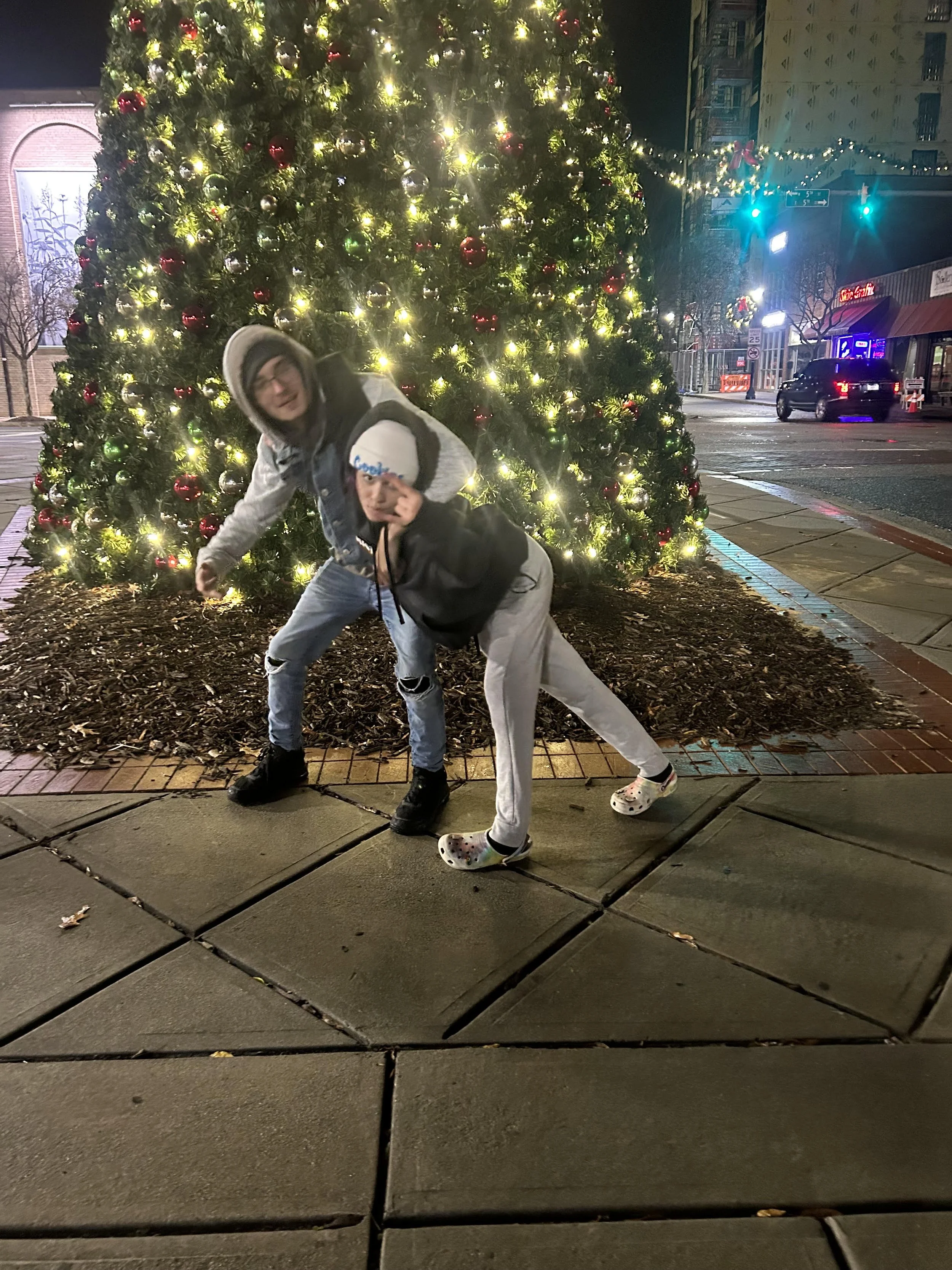 Makayla and her brother posing in front of a decorated Christmas tree at night on a city street with traffic lights and buildings in the background.