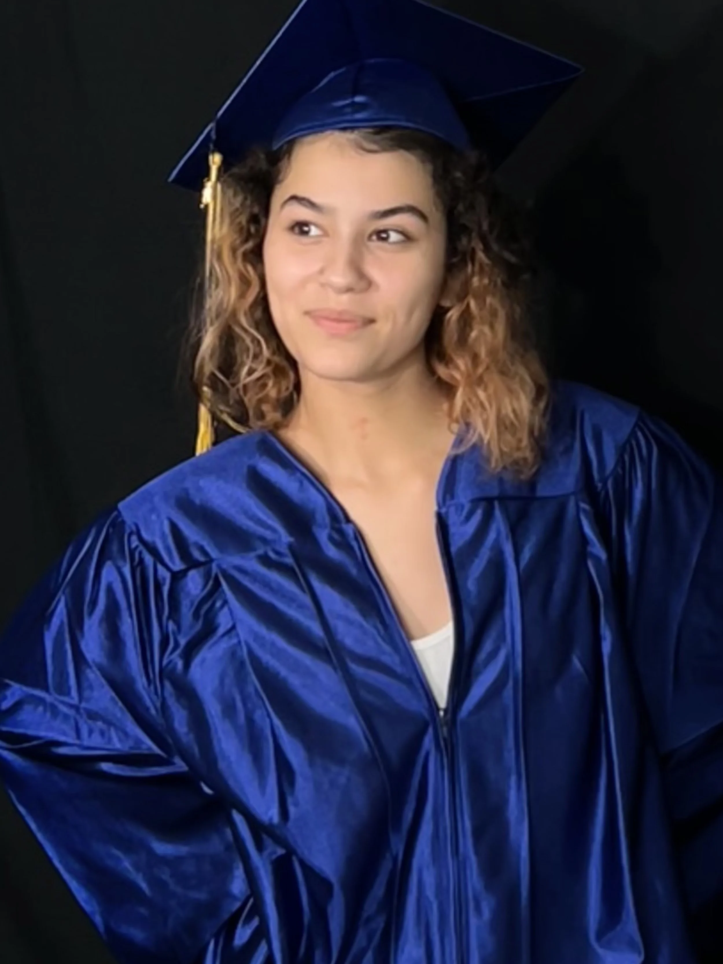 A young woman in a blue graduation gown and cap with a gold tassel, standing against a dark background.