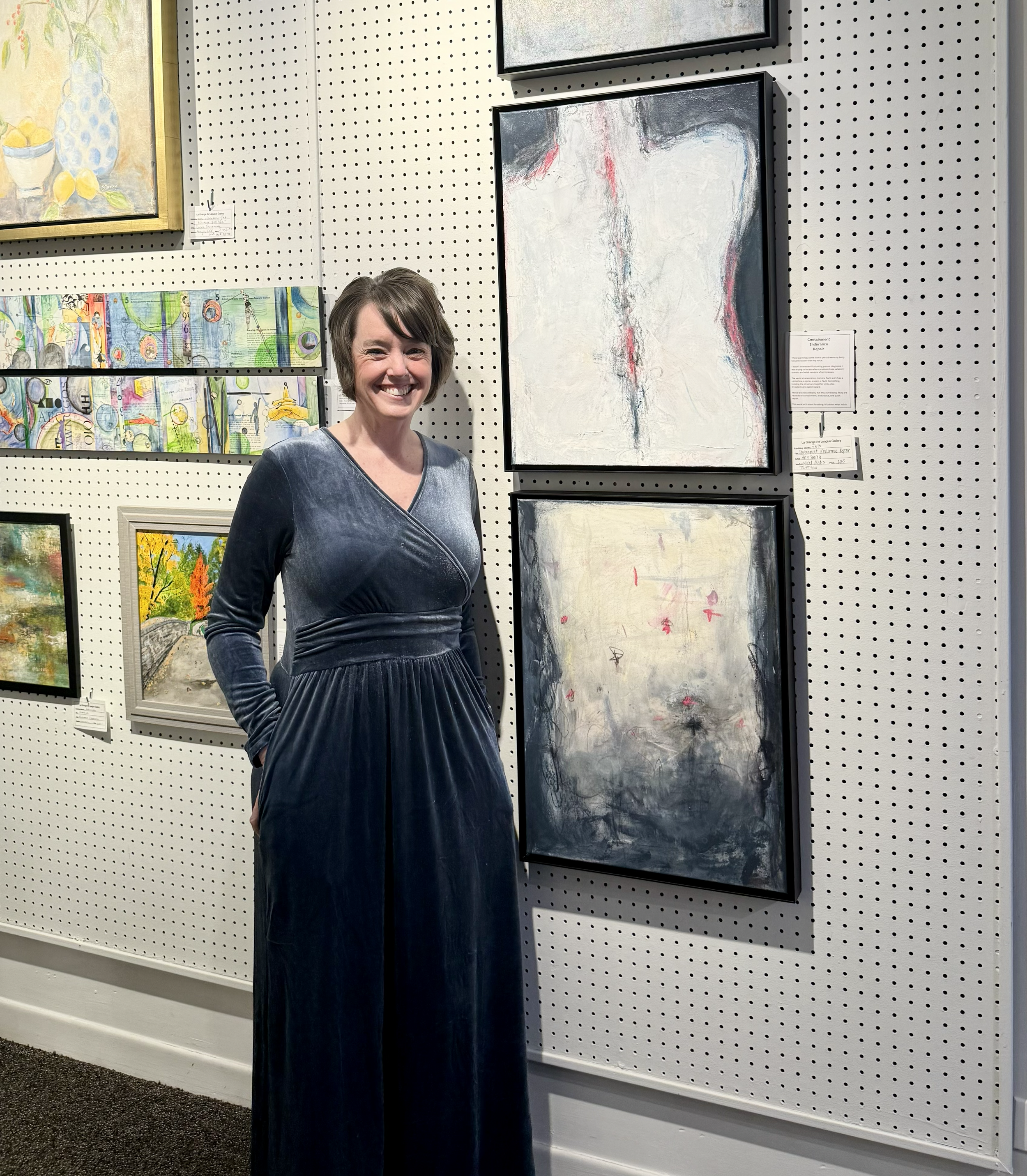 A woman with short brown hair smiling at an art gallery, standing in front of a wall with various abstract paintings and framed artwork.
