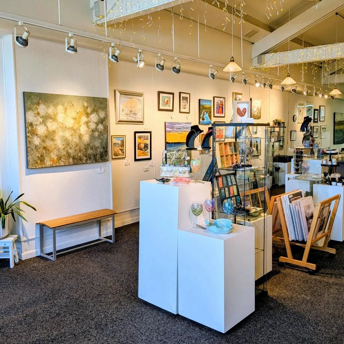 Interior of an art gallery with paintings, sculptures, and decorative items displayed on walls and shelves. Bright lighting and fairy lights hang from the ceiling.