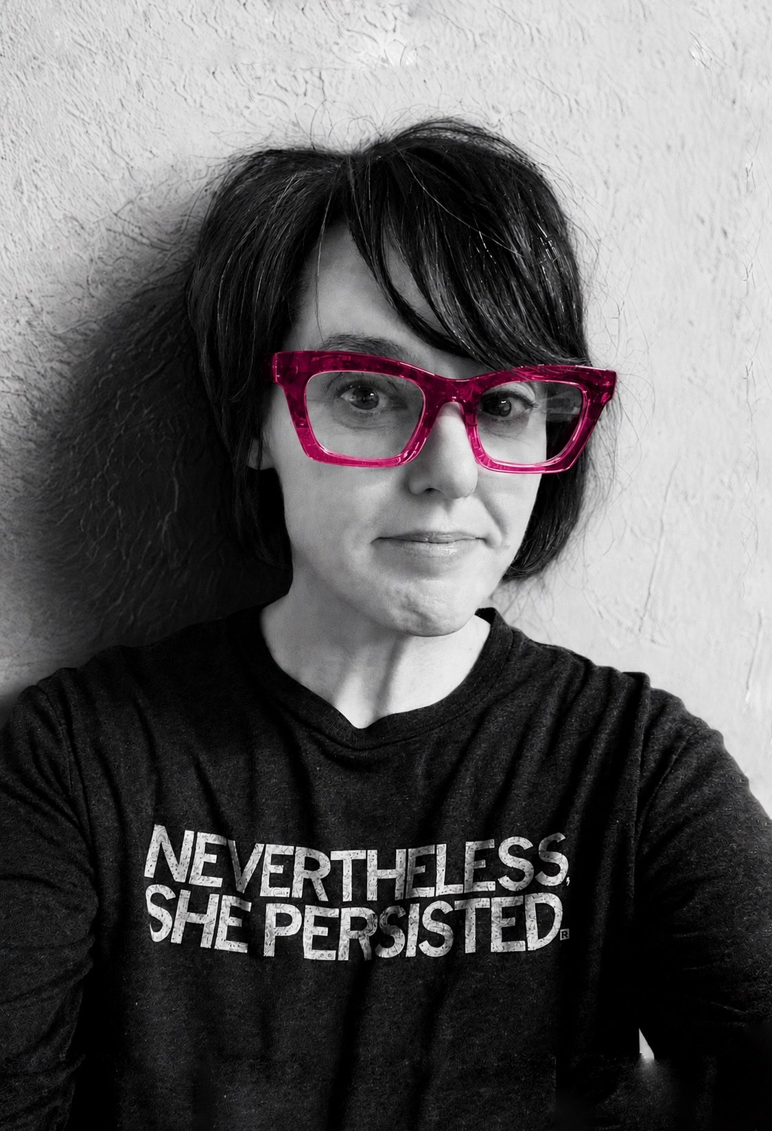 A woman with dark hair wearing large pink glasses and a black T-shirt with white text, standing against a textured wall.