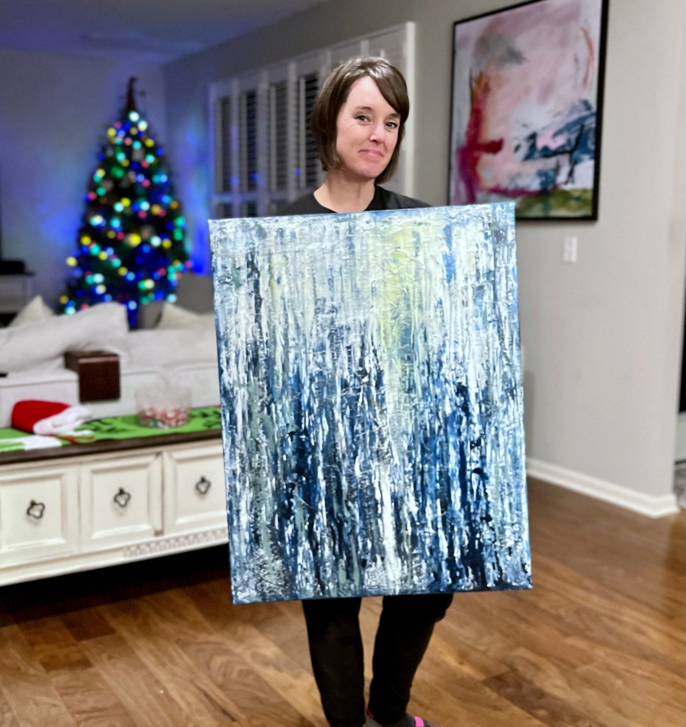 Person holding a shiny, reflective, abstract painting with a colorful cityscape background at home.