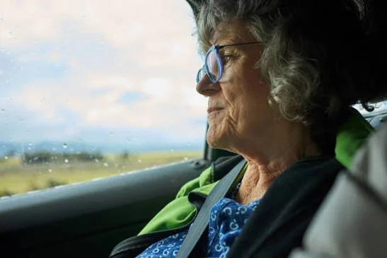 Older woman with glasses sleeping in the passenger seat of a car, looking out the window at a cloudy sky and open landscape.