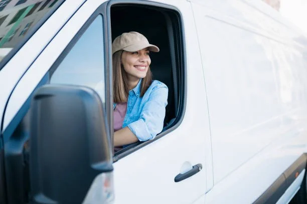 A woman smiling and looking out the window of a white delivery van, wearing a beige cap and a blue shirt.