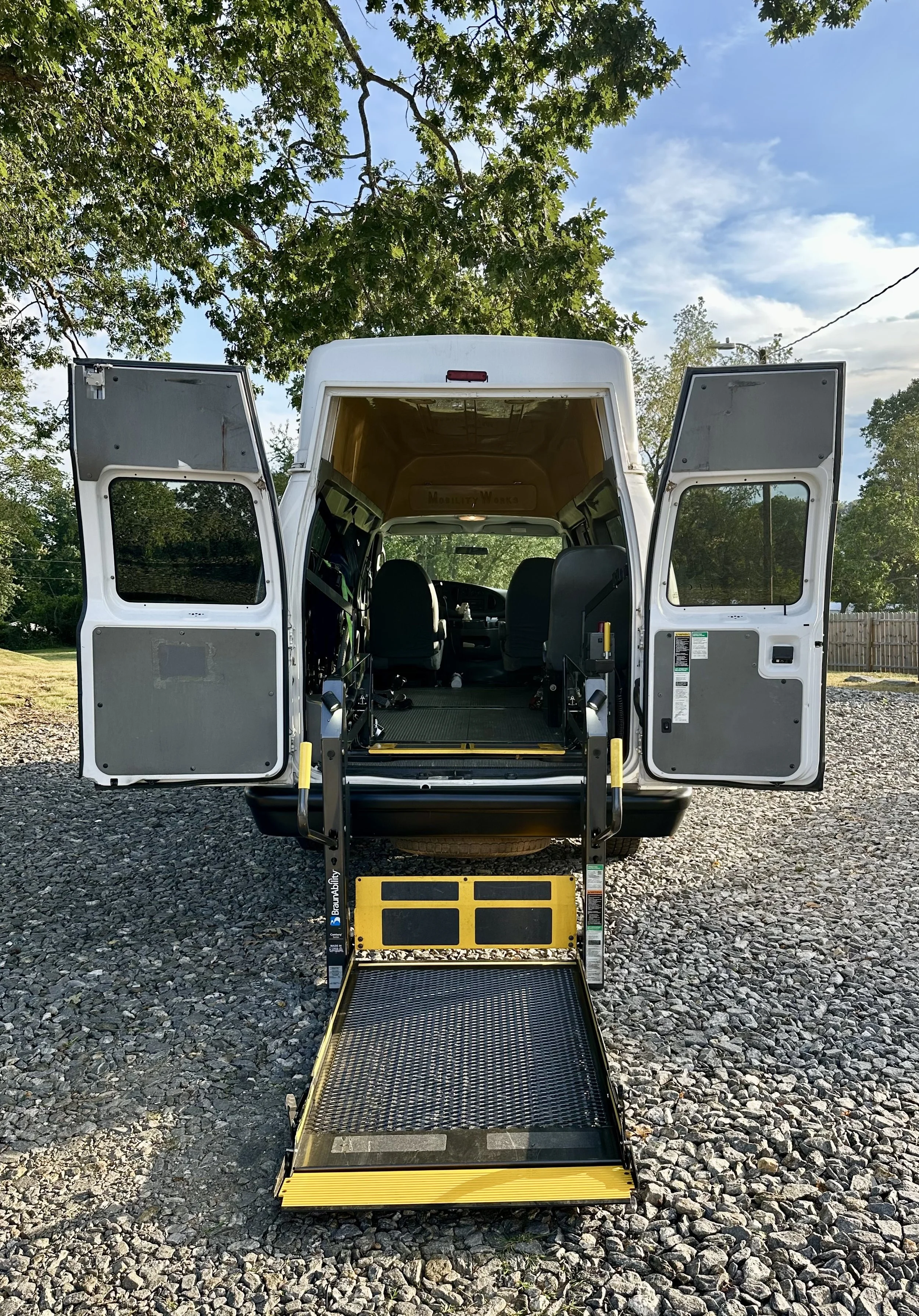 A white van with its rear doors open, showing a wheelchair lift at the back, parked on a gravel surface under a tree with green leaves and a blue sky with clouds.
