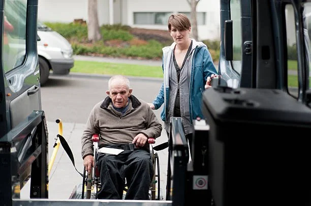An elderly man in a wheelchair being assisted by a younger woman near a vehicle.