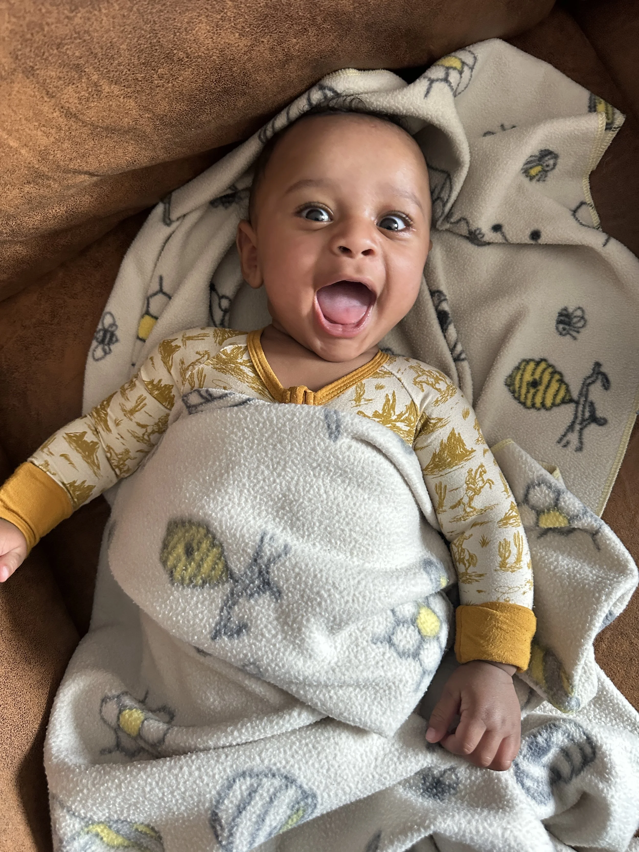 A baby with wide eyes and an open mouth, wrapped in a blanket with a bee pattern, lying on a brown couch.