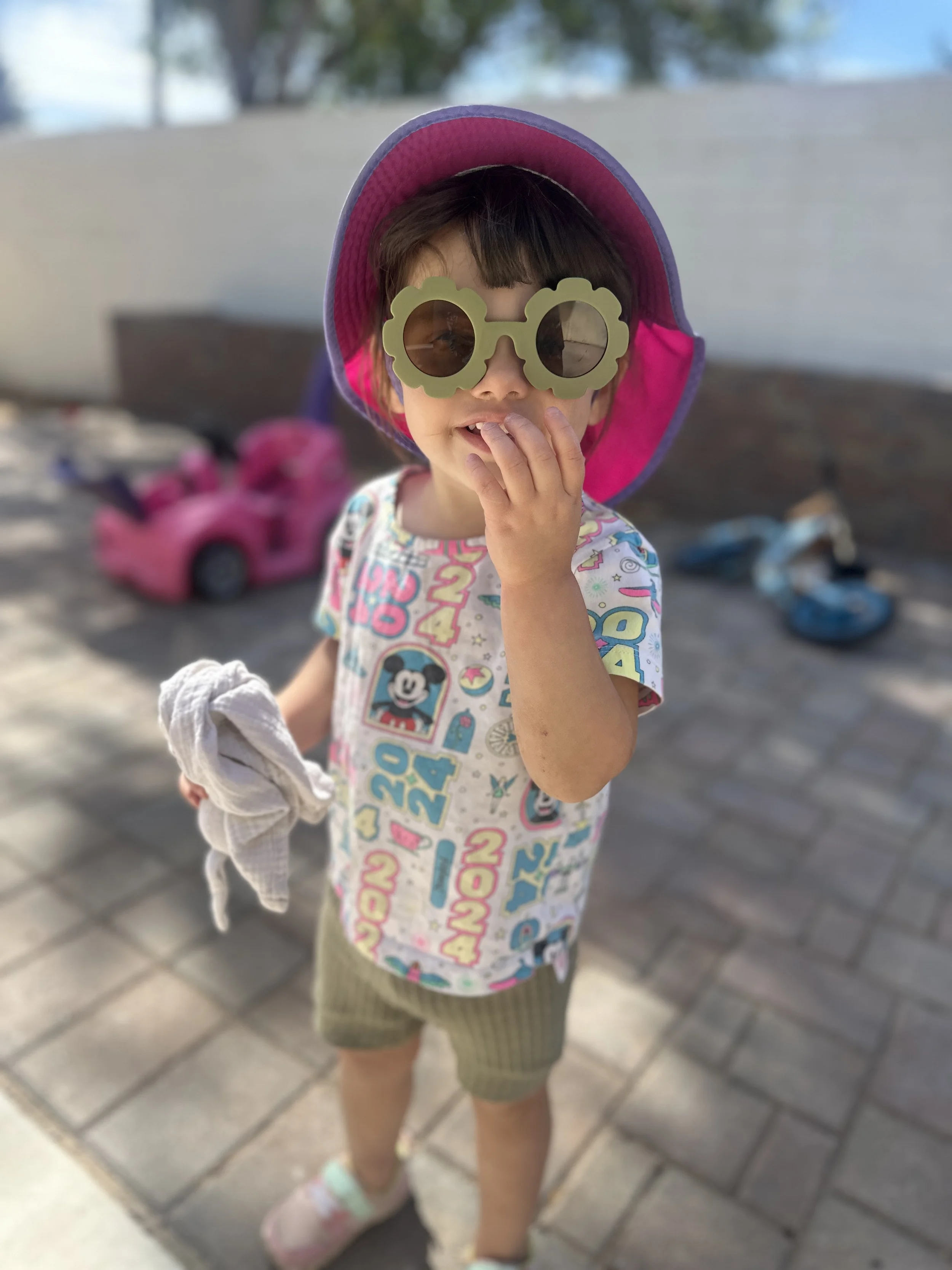 A young child wearing a purple hat, flower-shaped sunglasses, a colorful shirt with various characters, green shorts, and pink shoes, standing outdoors on a paved surface with toy cars in the background.