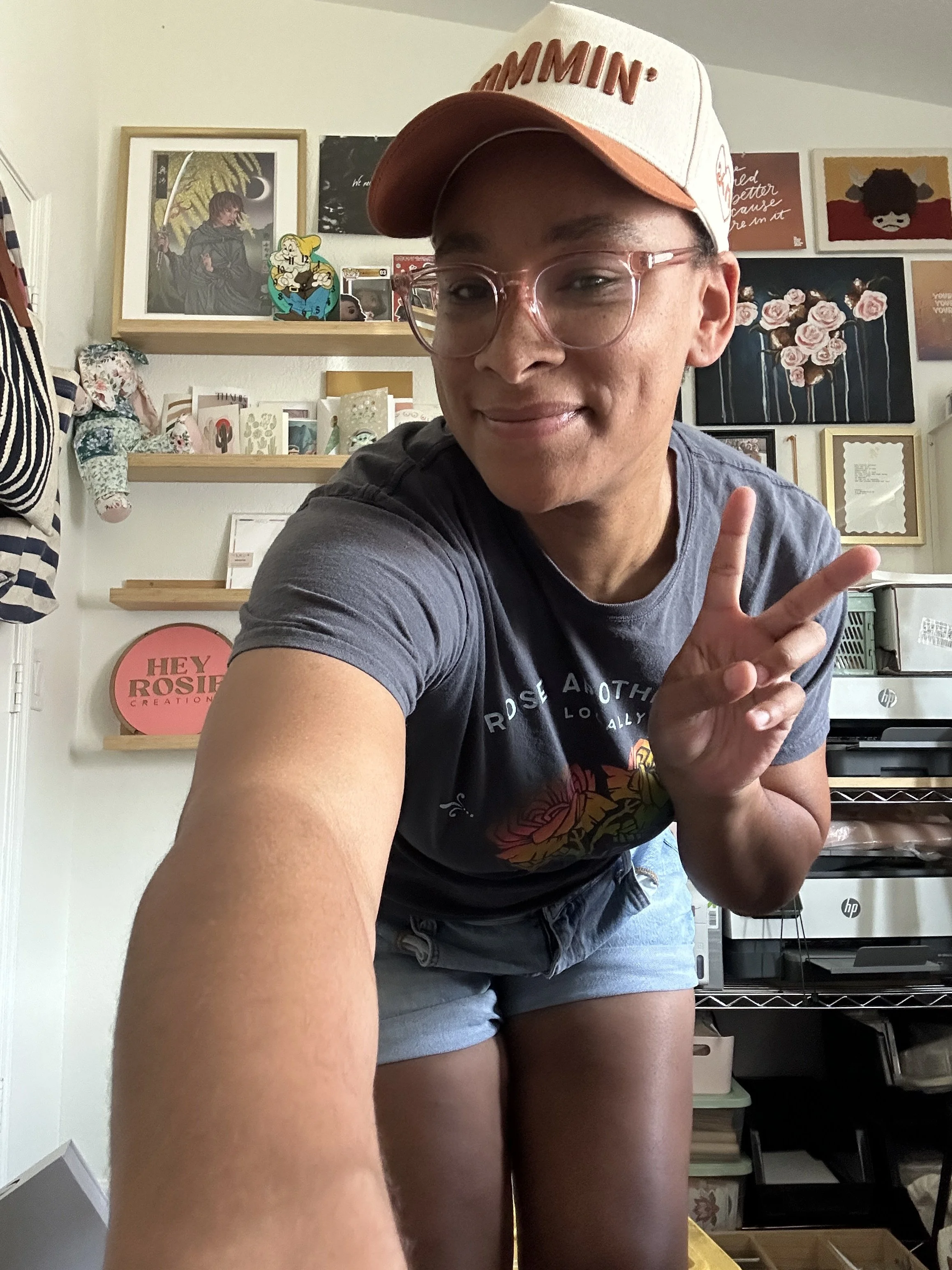 A person taking a selfie in a room filled with various artwork, books, and decorations on the shelves and walls. The person is making a peace sign with their hand, wearing glasses and a hat with the word 'HAMMIN' on it.