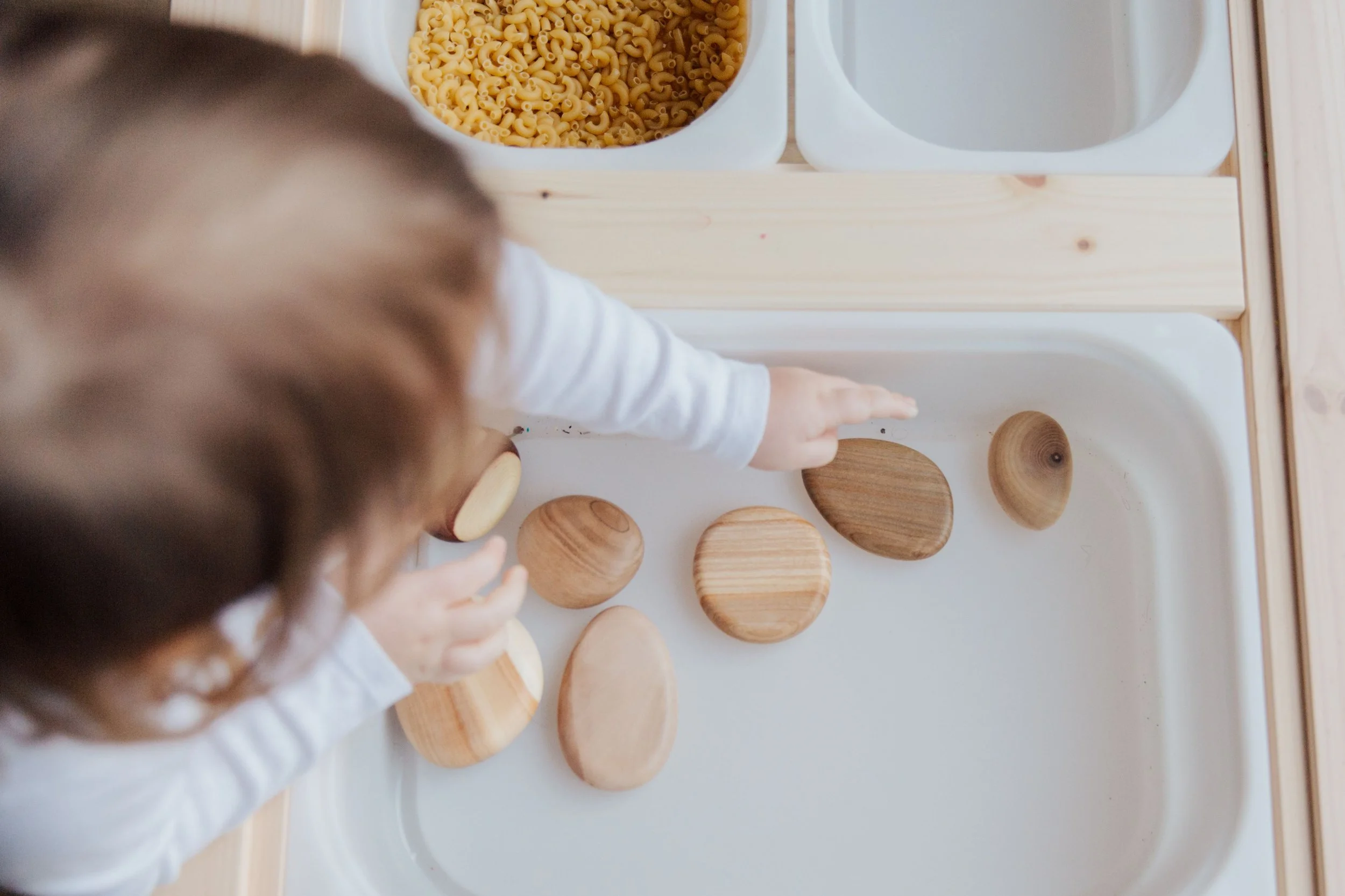 A child reaching out to touch wooden shape sorter toys on a white table, with two white bowls containing small pasta and an empty bowl in the background.