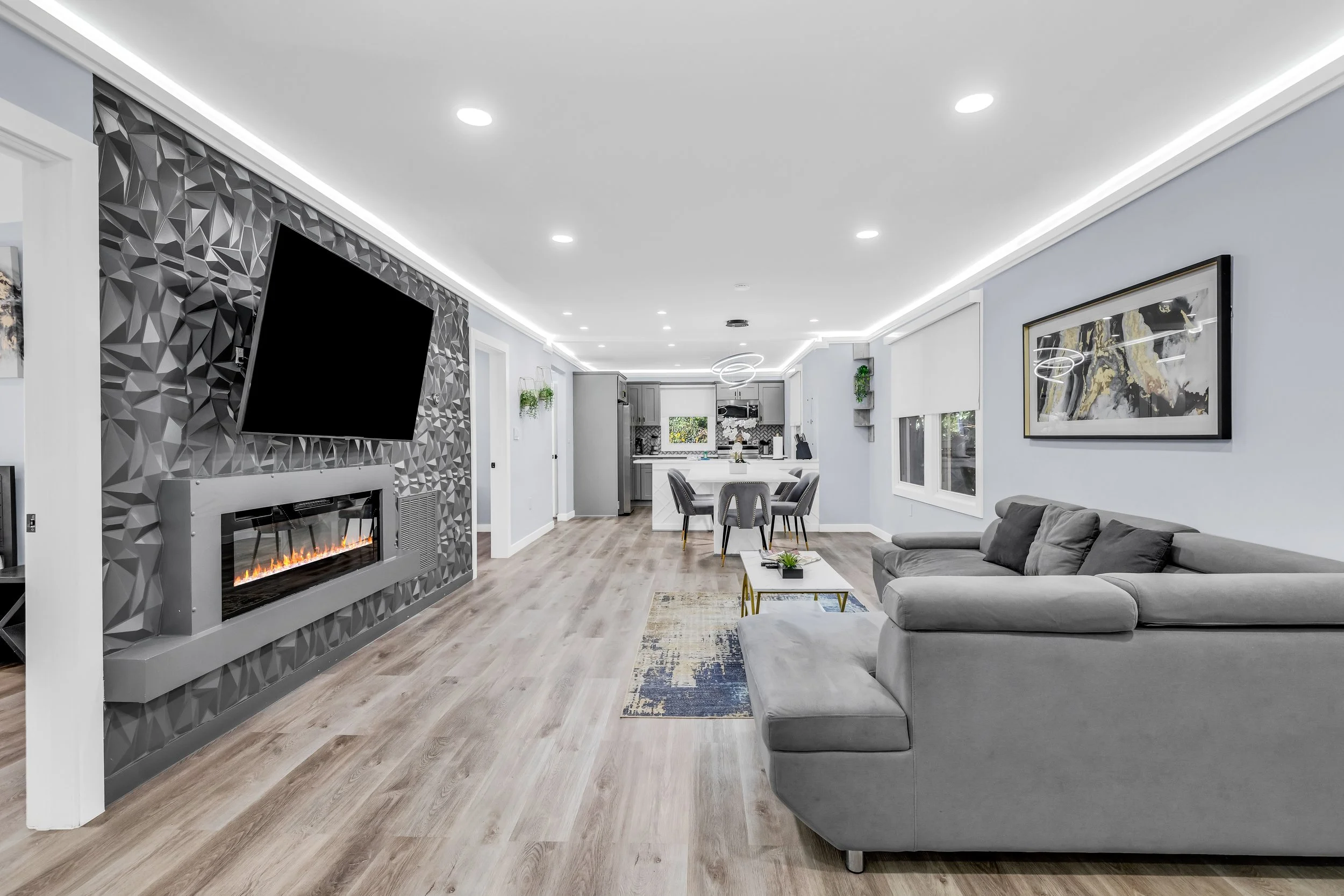 Modern living room with a gray sectional sofa, patterned rug, wall art, flat-screen TV, fireplace, and kitchen in the background.