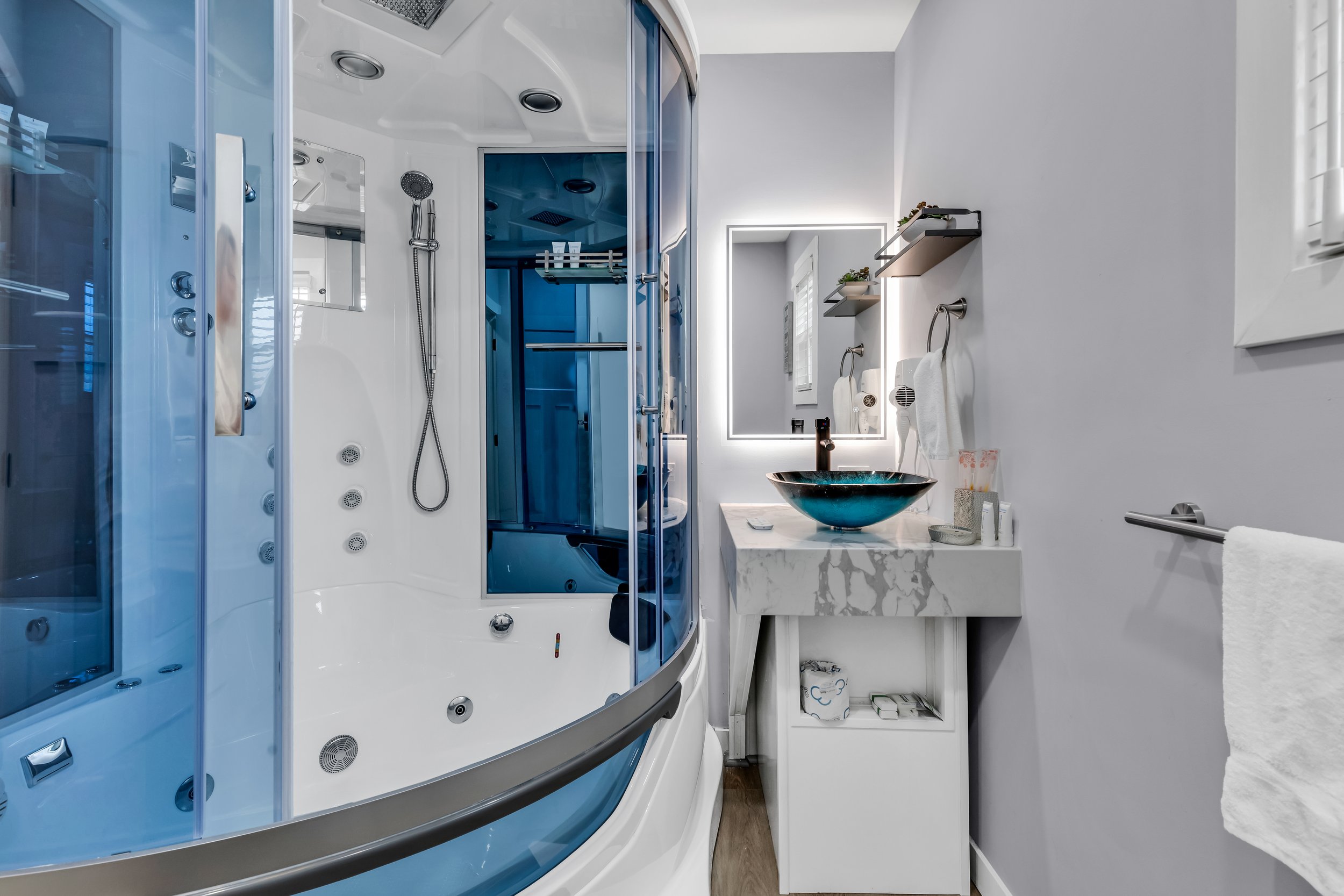 A modern bathroom featuring a blue and white small shower cabin with glass doors, a marble countertop with a black vessel sink, a large mirror with backlighting, and wall-mounted shelves with decorative plants. There are towels hanging on the wall, a small window, and various toiletries on the counter.