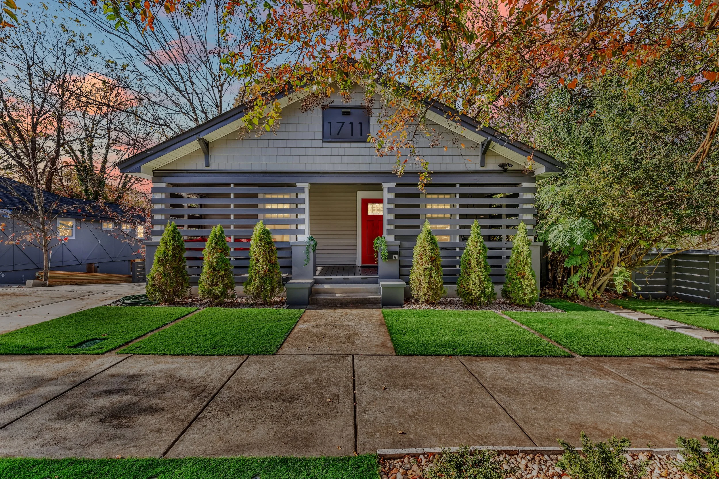 Front view of a modern house with a gray exterior, red front door, and a small porch with steps. There are small, neatly trimmed bushes in front and a concrete pathway leading to the entrance. The house is surrounded by trees with autumn-colored leaves.