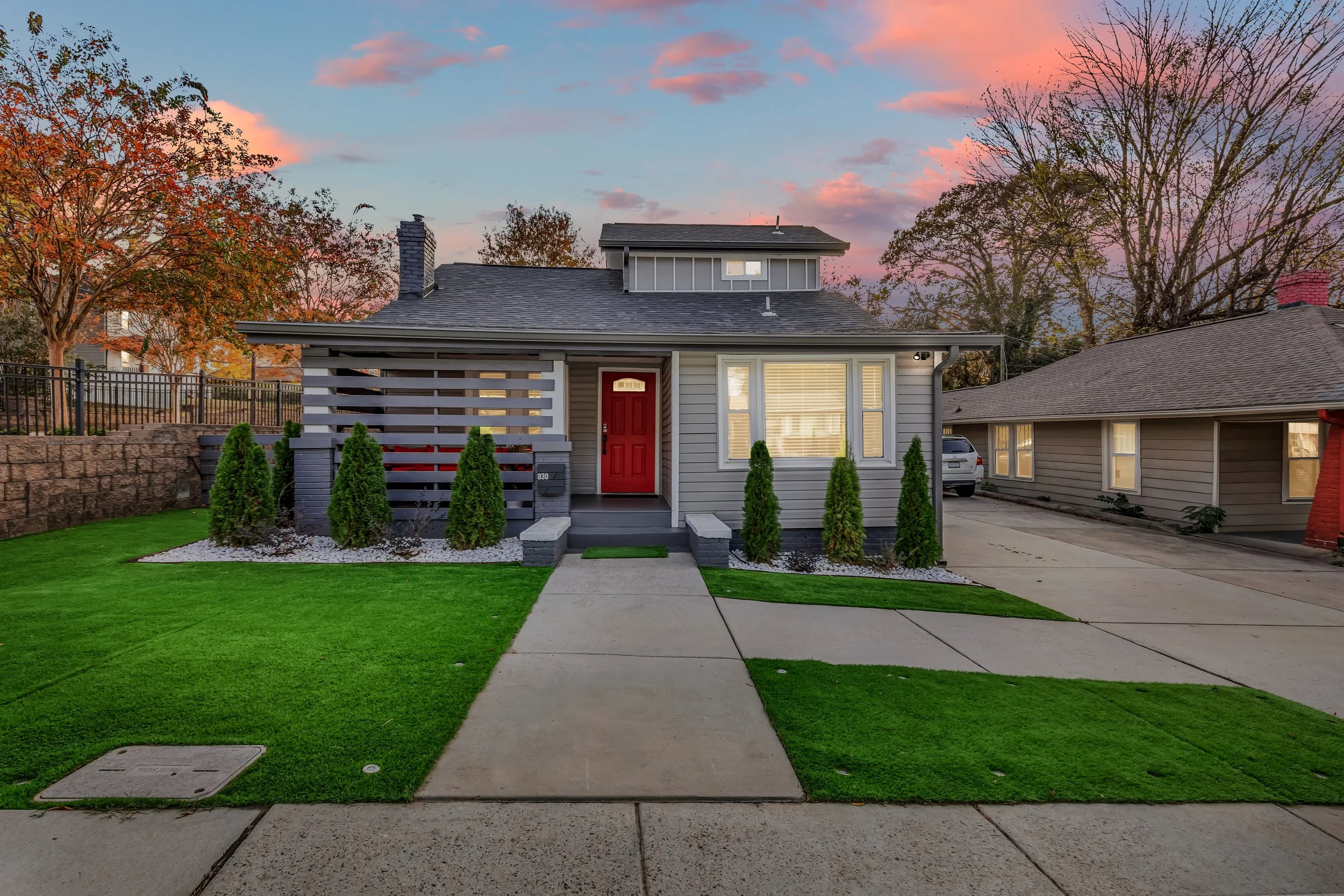 Front view of a modern house with a red front door, well-manicured lawn, small shrubs, concrete pathway, and neighboring houses under a colorful sky at sunset.