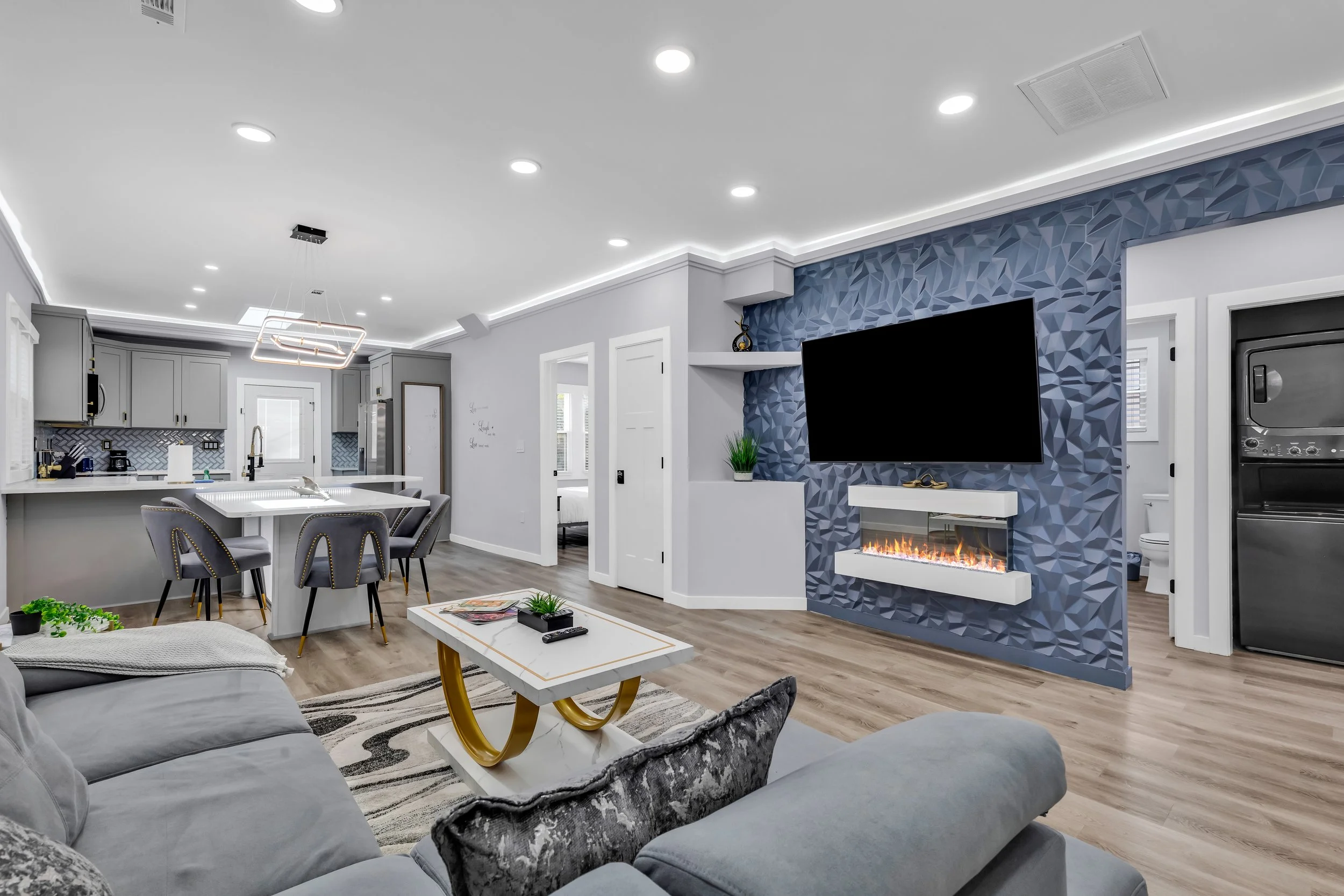 Modern living room with gray sofas, a white and gold coffee table, a flat-screen TV on a textured blue accent wall, and a kitchen with gray cabinets and a geometric backsplash.