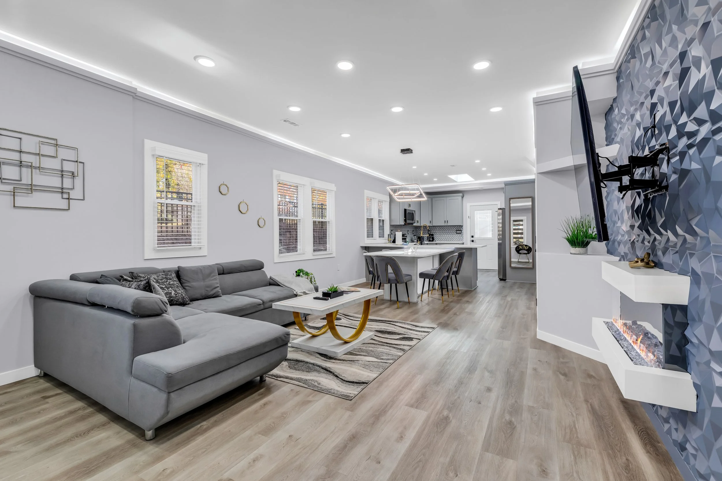 Modern open-concept living room and kitchen with gray sofa, abstract wall art, multiple windows, and a decorative fireplace with geometric blue tiles.