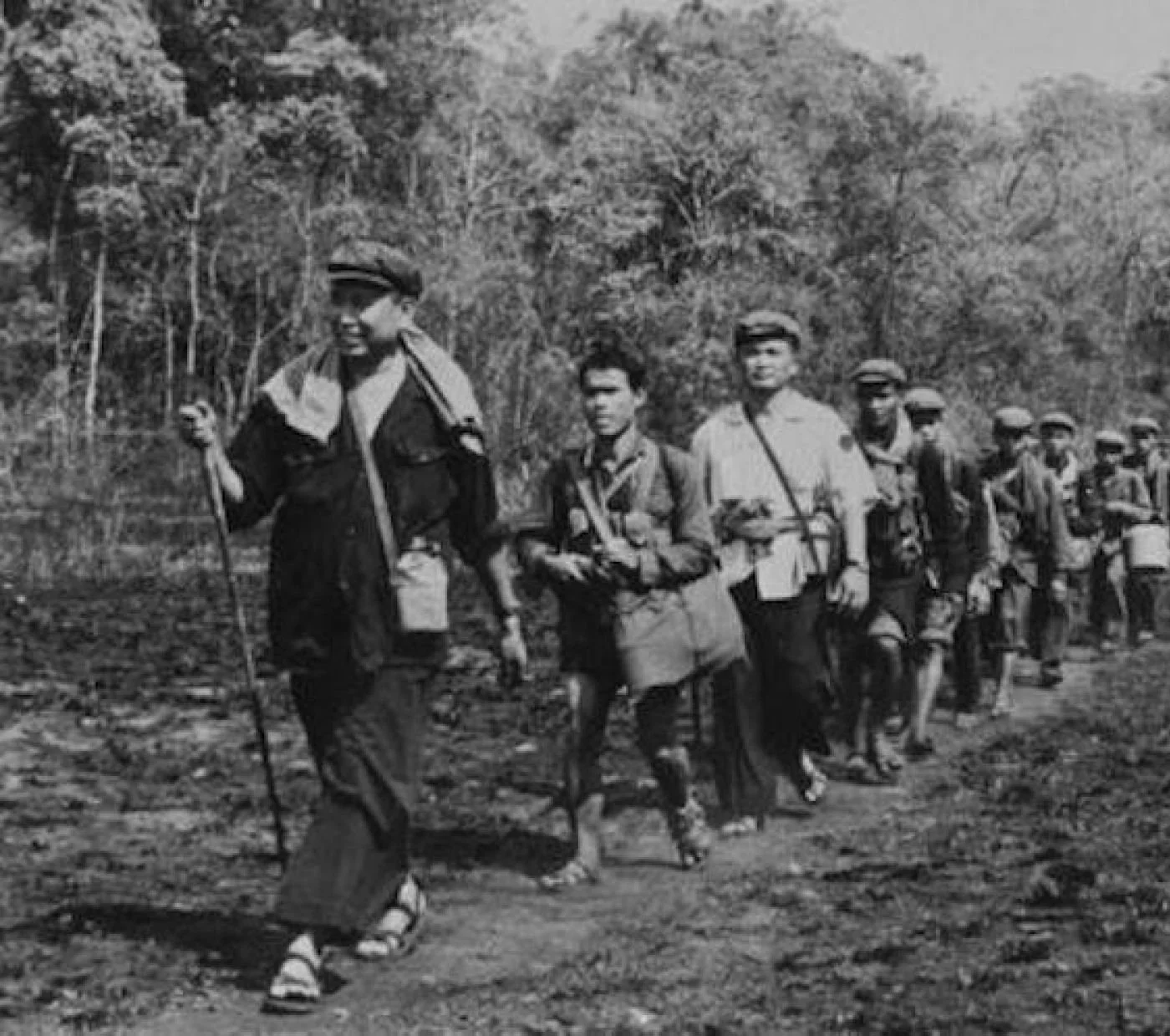 Line of children walking outdoors in a line, wearing caps and carrying bags, in a forested area.