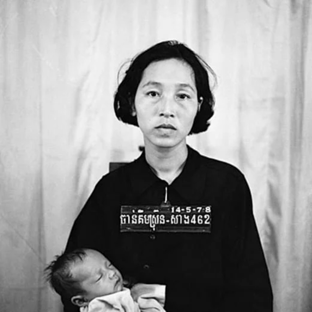 A black and white photo of a woman with short black hair holding a sleeping baby, with a plain background and a name tag or identification board displaying numbers and text.