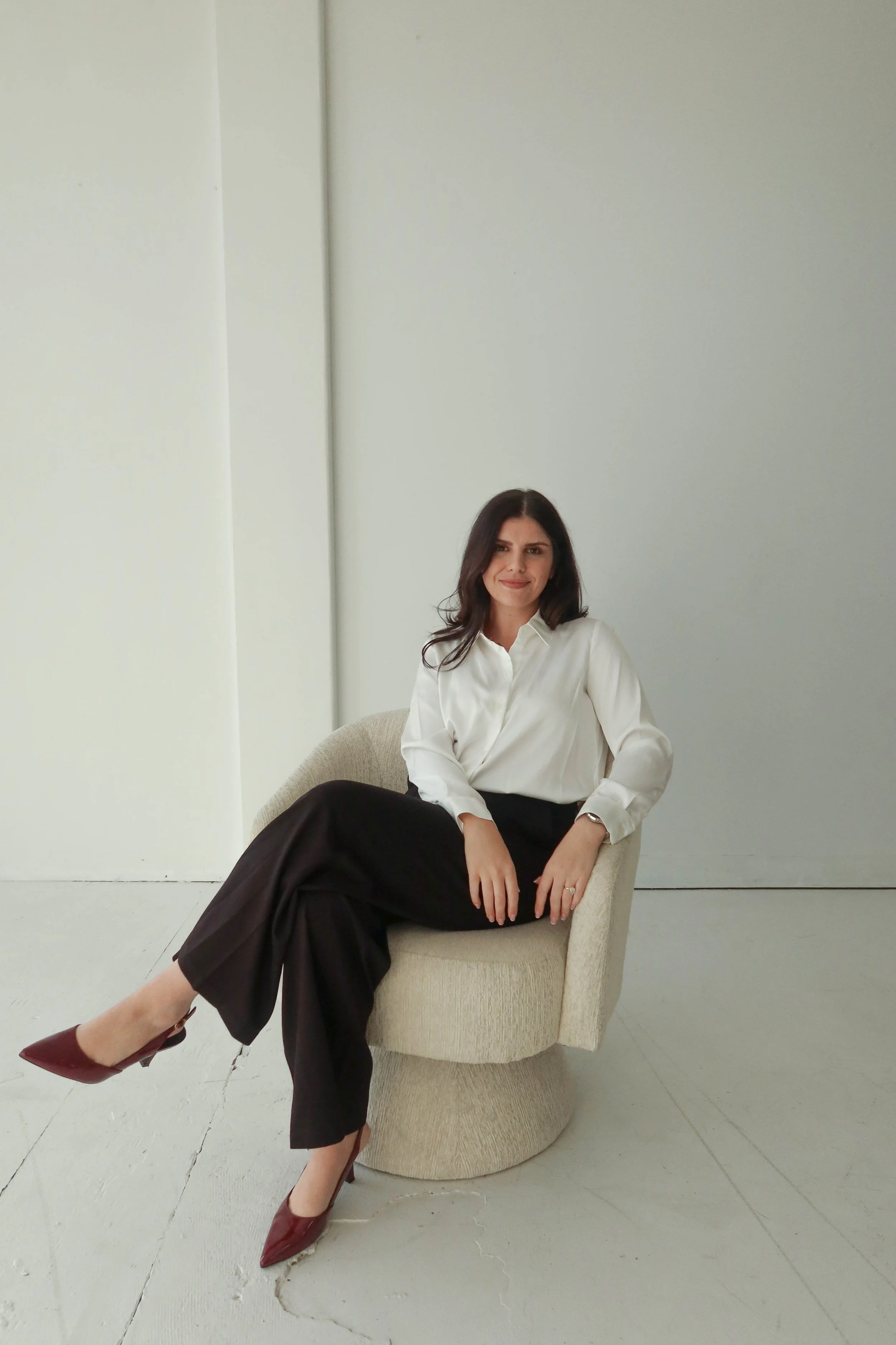 A woman with dark hair wearing a white shirt and black pants sitting in a beige armchair against plain white and light gray walls