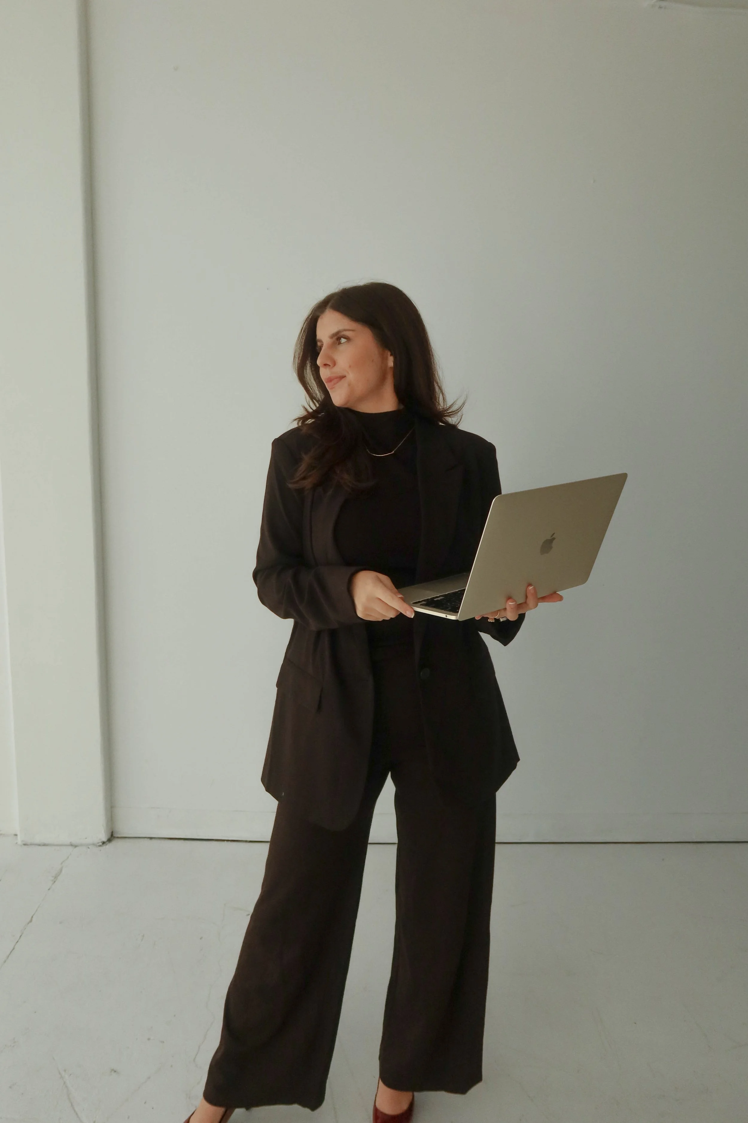 A woman with dark hair, dressed in a black blazer and black wide-leg pants, holding a silver laptop, standing against a plain white wall.