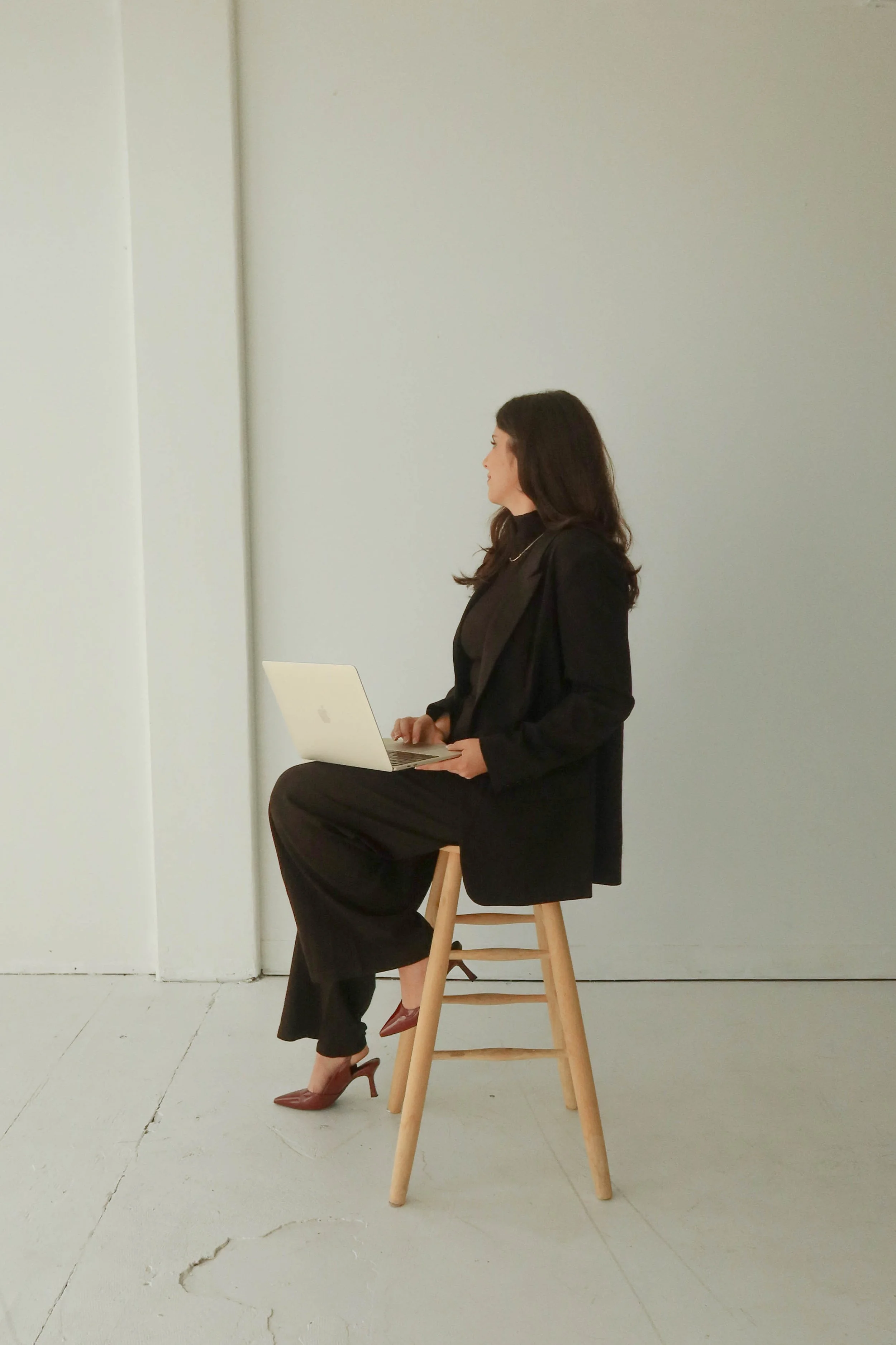 A woman with long brown hair is sitting on a wooden stool in front of a plain, light gray background. She is wearing a black blazer, black pants, and brown high-heeled shoes. She is looking to her right, at a silver laptop placed on her lap.